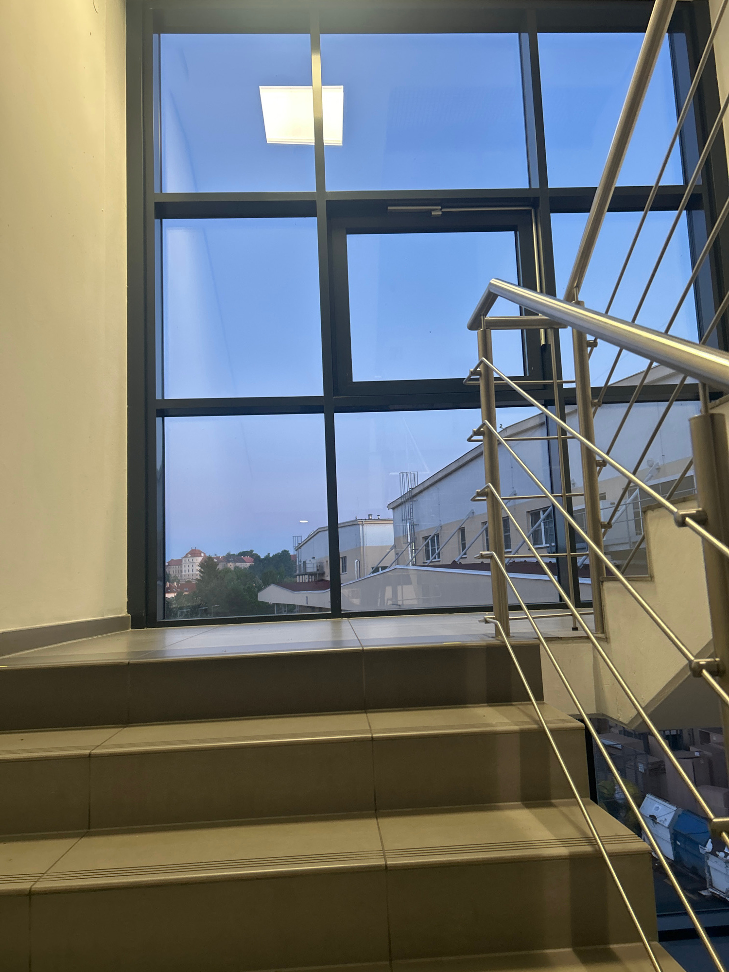The image shows a view looking down a staircase with a large window to the left. The staircase has tan-colored treads and risers, with a polished metal handrail running along the right side. The window is divided into multiple panes by dark metal frames. Through the window, a bright blue sky is visible, suggesting it is daytime. In the distance, a cityscape with buildings and trees can be seen, some of which appear to be old architecture. A single rectangular light fixture is visible on the wall to the left, inside the building. The overall scene is an interior view of a modern building, likely a public space or office, with a view of the outdoors. The image is taken from a high angle, looking downwards, implying the photographer is on an upper level of the staircase.