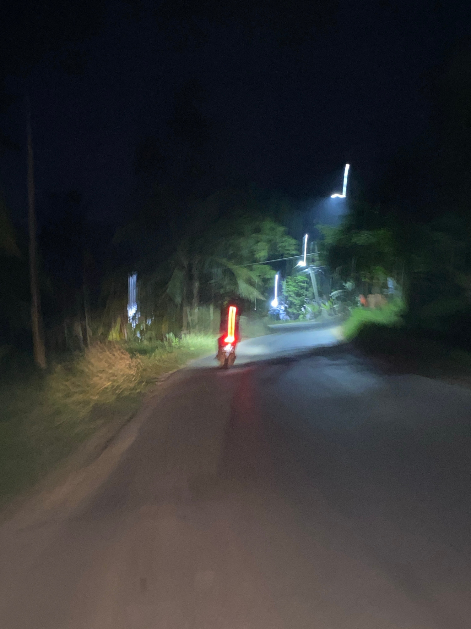 A nighttime, slightly blurry, medium shot captures a motorcycle driving on a paved road in Lhokseumawe, Indonesia. The road is dark and winding, with vegetation and palm trees lining the sides. The motorcycle, a prominent red object in the foreground, has a bright, elongated vertical taillight. Its headlights illuminate the road ahead, creating a bright beam that extends into the distance. In the background, there are more lights, some appearing as streaks of white and blue, suggesting streetlights or other illuminated structures. The overall atmosphere is dark and mysterious, with the motorcycle's lights cutting through the night.