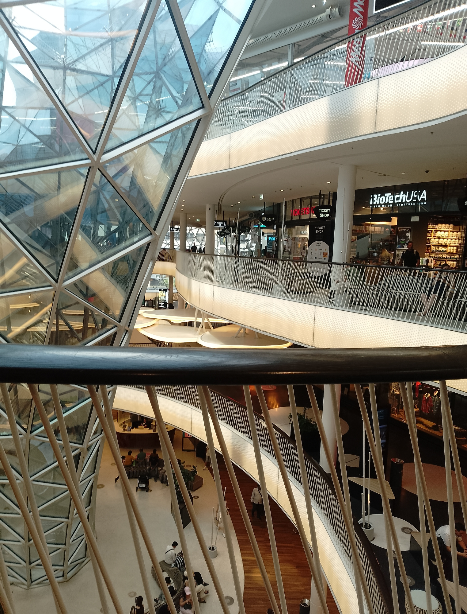 This is a medium-angle, eye-level shot captured from an elevated vantage point within a modern, multi-level shopping mall. The foreground is dominated by a dark wooden railing and a series of diagonally aligned, light-colored wooden poles that create a textured, semi-transparent barrier.
Beyond the foreground, the architecture of the mall unfolds. A striking, geodesic-like glass structure with a white frame is visible on the left, reflecting the interior lights and other levels of the mall. Multiple levels of walkways with light-colored railings are connected by ramps and bridges, showcasing a contemporary design. On the upper levels, various shop storefronts are visible, including "BiotechUSA" and "TICKET SHOP." People are seen walking and standing on these different levels, some gathered in small groups. The flooring on the lower levels appears to be a combination of light-colored tiles and a warm-toned wood. The lighting is bright and diffuse, typical of an indoor commercial space. The scene depicts a busy, well-lit commercial environment.