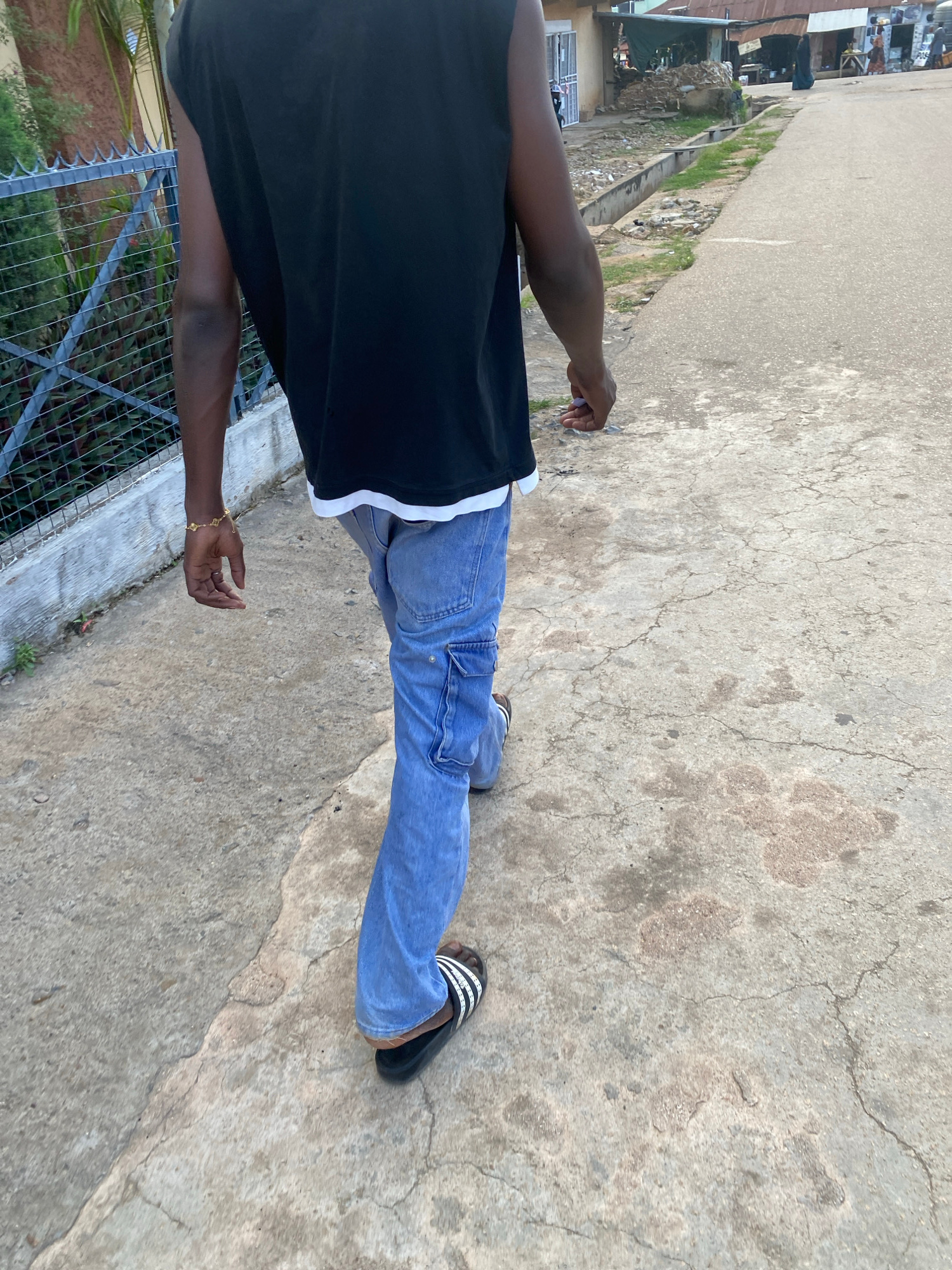 A person is captured from a slightly elevated, sideways perspective, facing away from the camera. They are wearing a black t-shirt with a white stripe on the neckline, blue denim jeans with cargo pockets, and black and white striped slides. The person is standing on a cracked concrete surface, with a paved road and some vegetation visible in the background. On the left side of the frame, there's a metal fence and some foliage. The lighting suggests it is daytime, possibly late afternoon given the soft shadows. The scene appears to be outdoors, in a casual urban or semi-urban environment, potentially in Jos, Nigeria, as indicated by the location context. No specific activity is clearly depicted, but the pose suggests the person might be walking or standing still, possibly observing something. There is no visible text in the image. The overall mood is neutral.
