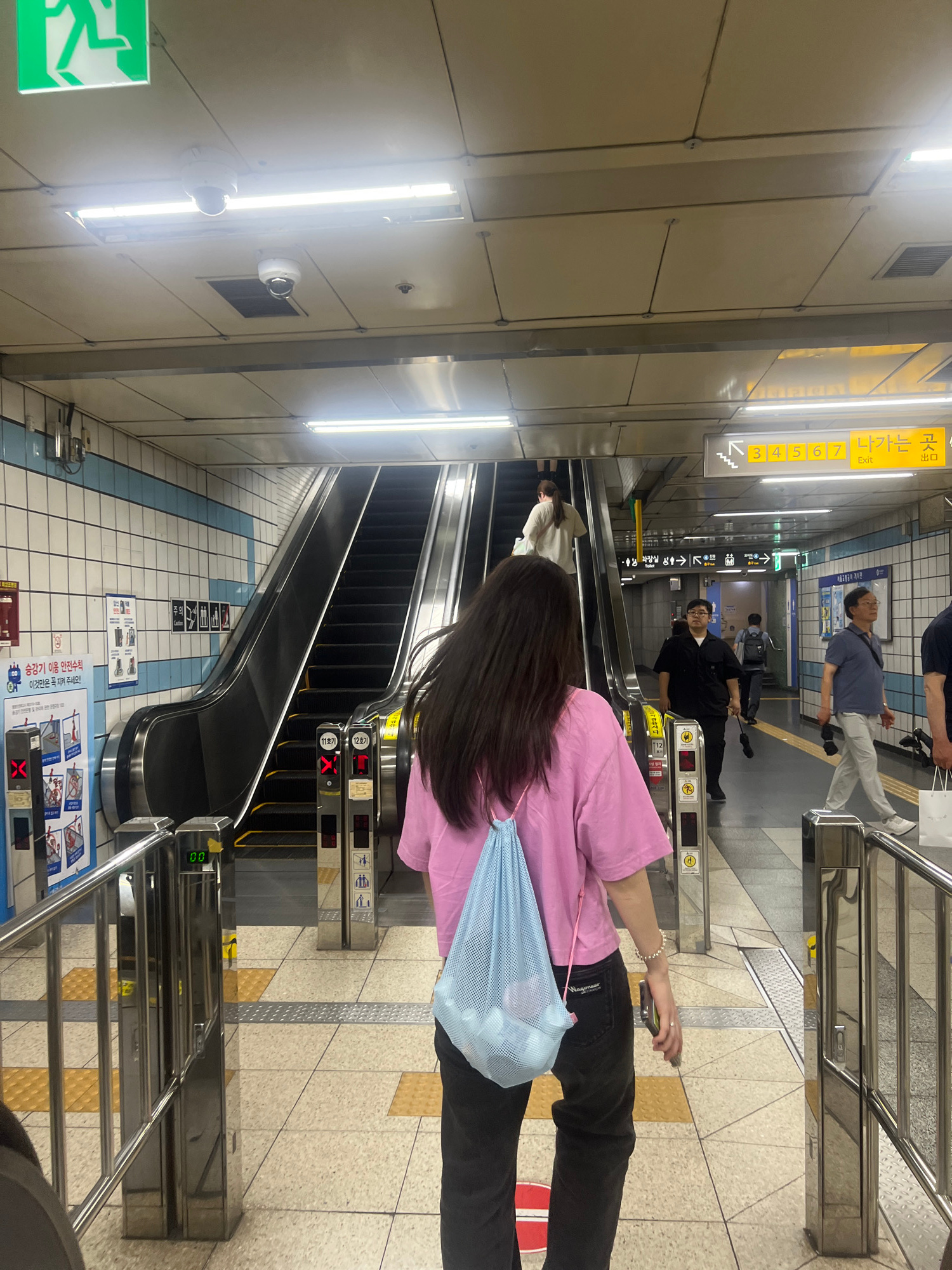 The image depicts a scene inside a subway station in Seoul, South Korea. The perspective is from behind a young woman wearing a pink t-shirt and black pants, carrying a light blue mesh bag over her shoulder. She is walking away from the camera towards an escalator.
The environment is brightly lit with fluorescent lights on the ceiling. There are two escalators visible; one is moving upwards and has several people on it, including a woman in a white top. The other escalator is to the right and appears to be stationary, with turnstiles in the foreground.
Signs are visible throughout the station, including an "Exit" sign in Korean and English with directional arrows and numbers indicating exits 3, 4, 5, 6, and 7. Another sign on the wall displays safety instructions for escalator use. There are also security cameras mounted on the ceiling.
Other individuals are present in the background, including a man in a black shirt and glasses, a man in a blue shirt and grey pants, and another person with a backpack. The floor is tiled with a pattern of beige and yellow squares. Metal railings and turnstiles are prominent features of the station's infrastructure. The overall impression is of a busy public transportation hub.
