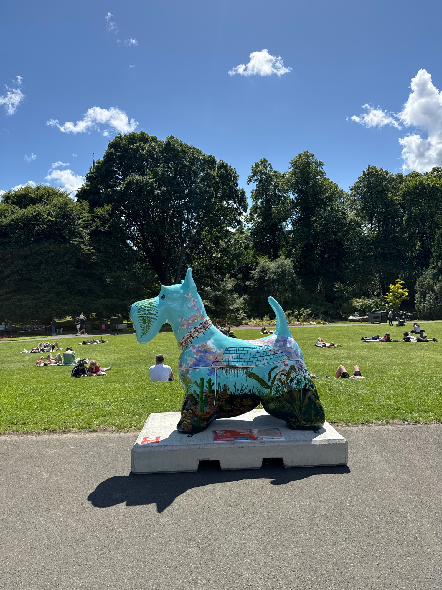 In the city of Glasgow, United Kingdom, a large, brightly painted sculpture of a Scottie dog stands on a concrete base in a park. The sculpture is predominantly turquoise with intricate designs featuring plants, animals, and buildings. A few people are visible in the background, scattered across the grassy park, many of whom are lying down or sitting on blankets, enjoying the sunny weather. The park is surrounded by mature trees, and the sky above is a clear blue with scattered white clouds. The sculpture's shadow is cast onto the paved path in the foreground.