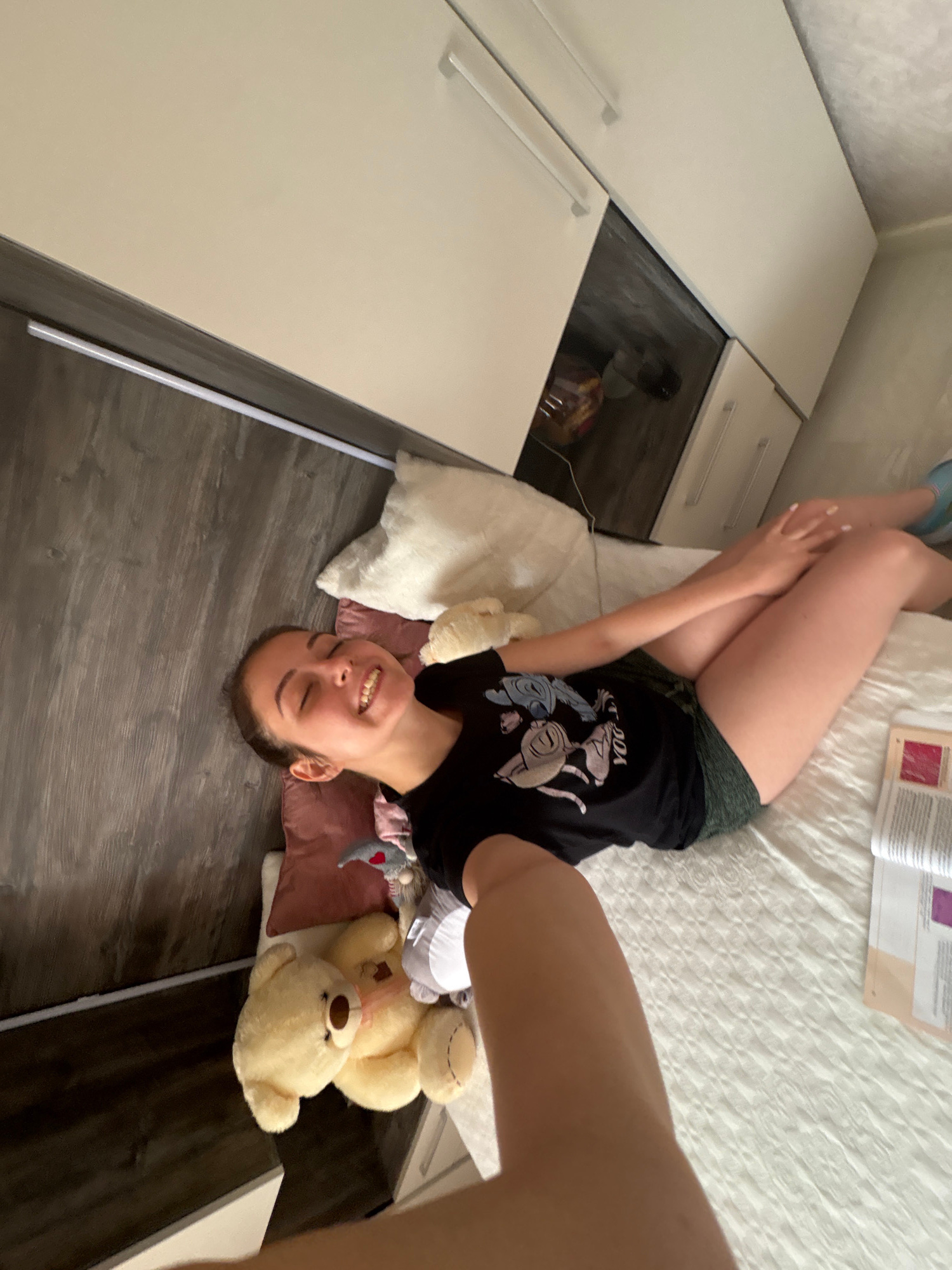 A young woman with dark hair is lying on a bed, holding up a camera to take a selfie. She is wearing a black t-shirt with a graphic print and green shorts. Her eyes are closed, and she is smiling. The bed is covered with a white quilted comforter. To the left of the woman is a large teddy bear and several pillows. In the background, there is a wardrobe with white doors and a dark wood-grained panel. The image appears to be taken indoors, possibly in a bedroom. The location is identified as Bulgarovo, Bulgaria.
