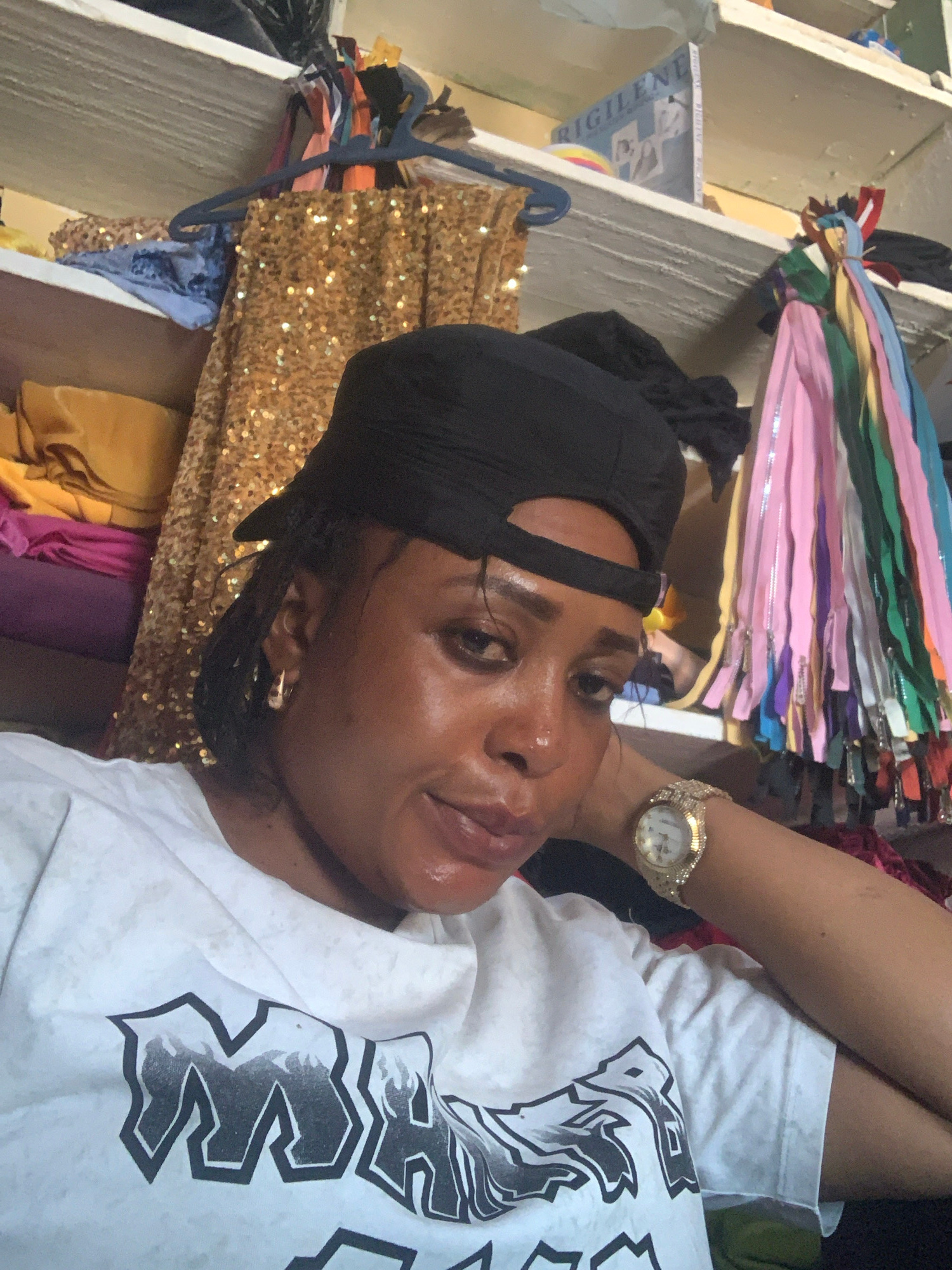 A person is lying down, looking up towards the camera. They are wearing a black baseball cap and a white t-shirt with a black graphic design. A gold watch with rhinestones is visible on their left wrist. The background appears to be a storage area or closet, with shelves filled with various items. Notably, there are stacks of colorful zippers in various shades of pink, blue, green, and yellow. A sequined gold garment is also visible hanging on a rack. The lighting suggests it could be daytime. The image is oriented vertically, with the subject's head towards the bottom and their body extending upwards. The overall impression is a casual, personal snapshot taken in a cluttered environment.