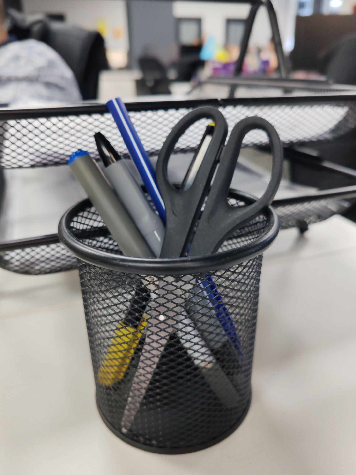 A close-up, eye-level, slightly angled shot captures a black mesh pen holder overflowing with stationery. Dominating the foreground, the pen holder sits on a white surface. Inside, a pair of black-handled scissors with visible finger loops is prominently displayed. Behind the scissors, a blue and white pen stands upright. Next to it, a grey pen is also visible. Further down, partially obscured, are other writing instruments including a yellow marker and a dark pen. The background is blurred, suggesting an office environment with cubicles and office chairs, but specific details are indistinct. There is no indication of the city of Poltava, Ukraine, or any other specific geographical location. The scene depicts a typical desk setup for work or study.