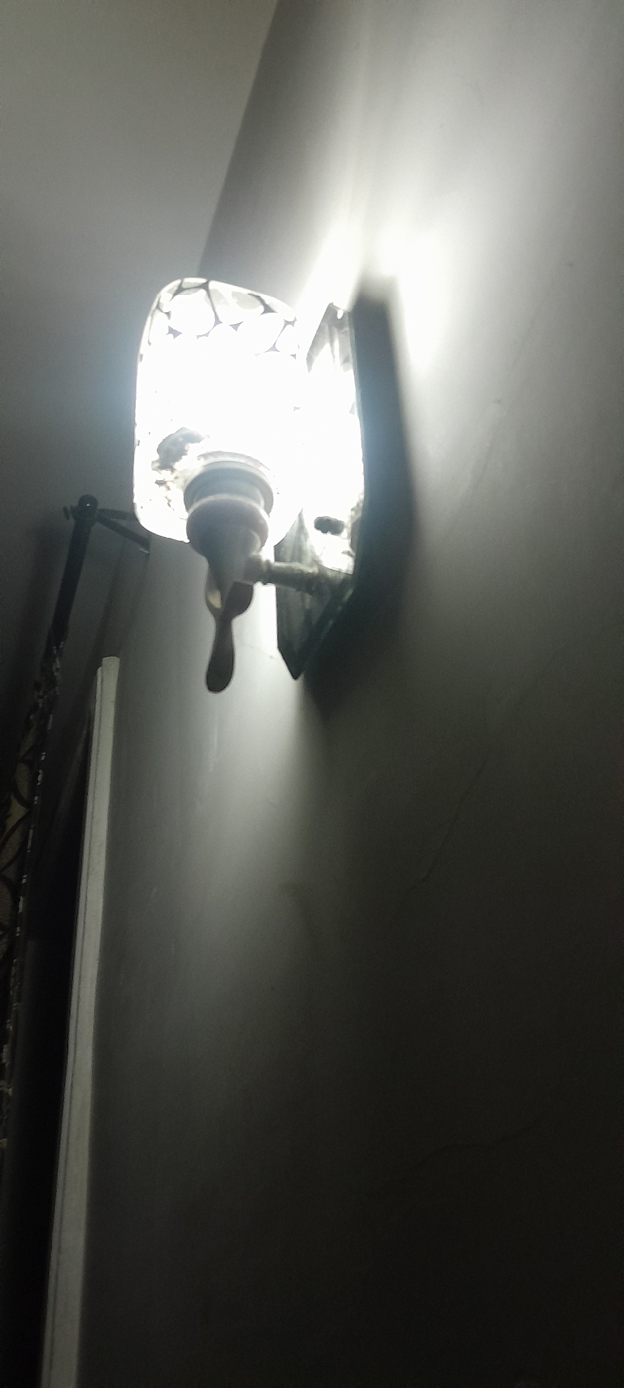 An outdoor wall lamp is mounted on a plain, light-colored wall, casting a bright light and a distinct shadow. The lamp has a decorative, possibly glass, shade with a patterned design. Below the shade, a fixture with a tap-like handle extends downwards. To the left of the lamp, a portion of a dark, decorative fence or railing is visible, along with a vertical white trim or frame. The scene is dimly lit, with the lamp being the primary light source. The overall environment appears to be an exterior wall in an unspecified urban location.