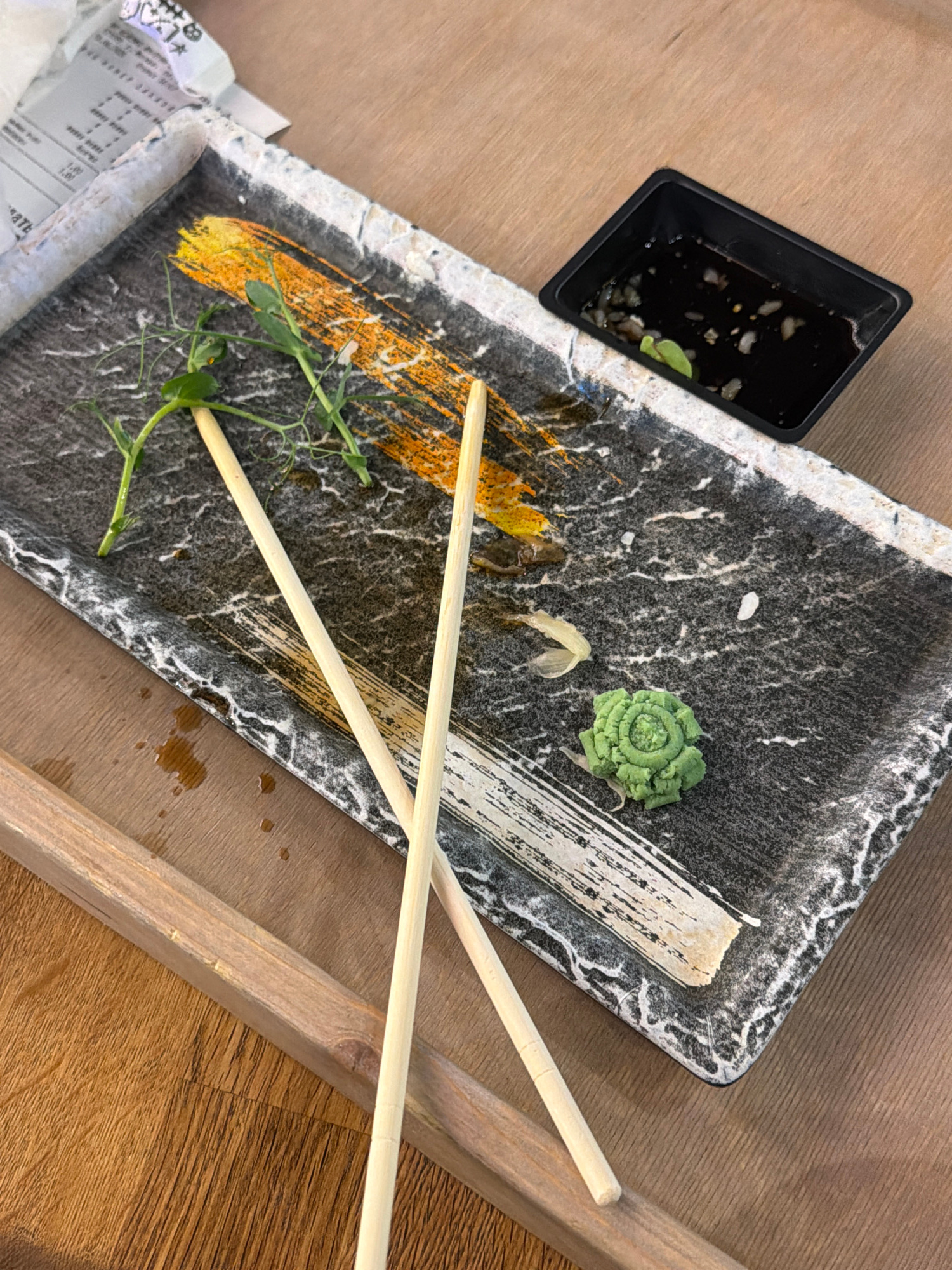 An overhead, close-up shot shows a dark gray, rectangular plate with a marbled design resting on a wooden surface. The plate contains two light-colored chopsticks, a small pile of bright green wasabi, a few delicate green sprigs, a smear of orange sauce, and a small black dish filled with dark liquid and some scattered food scraps. Part of a white paper receipt is visible in the upper left corner of the plate. A wooden plank runs across the bottom of the frame, obscuring the lower portion of the plate and the table. The lighting is moderate, and the scene appears to be set in an indoor dining establishment. The location is not specified in the image.