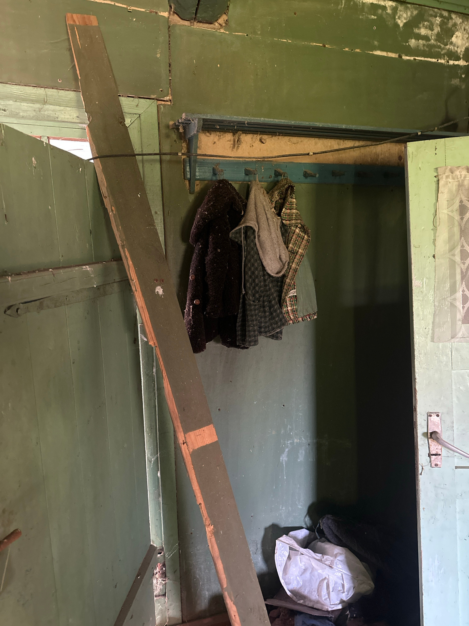 The image depicts a cluttered, dimly lit interior space, likely a storage area or attic. The dominant color is a faded green, applied to wooden walls and surfaces. A large wooden plank runs diagonally across the foreground, obscuring part of the scene.
In the middle ground, a pile of folded fabrics is visible, featuring checkered and plain dark patterns. To the right of the fabrics, a white plastic bag and some dark clothing are stuffed into a dark opening, suggesting it's a cabinet or cubby. A thin black cable extends from the top left, running downwards and secured with small metal clasps to the green wall.
The setting appears weathered and somewhat neglected, with peeling paint and visible nails on the wooden surfaces. A lace curtain is visible at the top of the opening, partially obscuring the interior. The lighting suggests it might be daytime, but the lack of direct sunlight and the deep shadows create a somber atmosphere. There is no visible text in the image. The overall impression is one of disuse and accumulated items in a rustic, possibly rural, Belarusian interior.