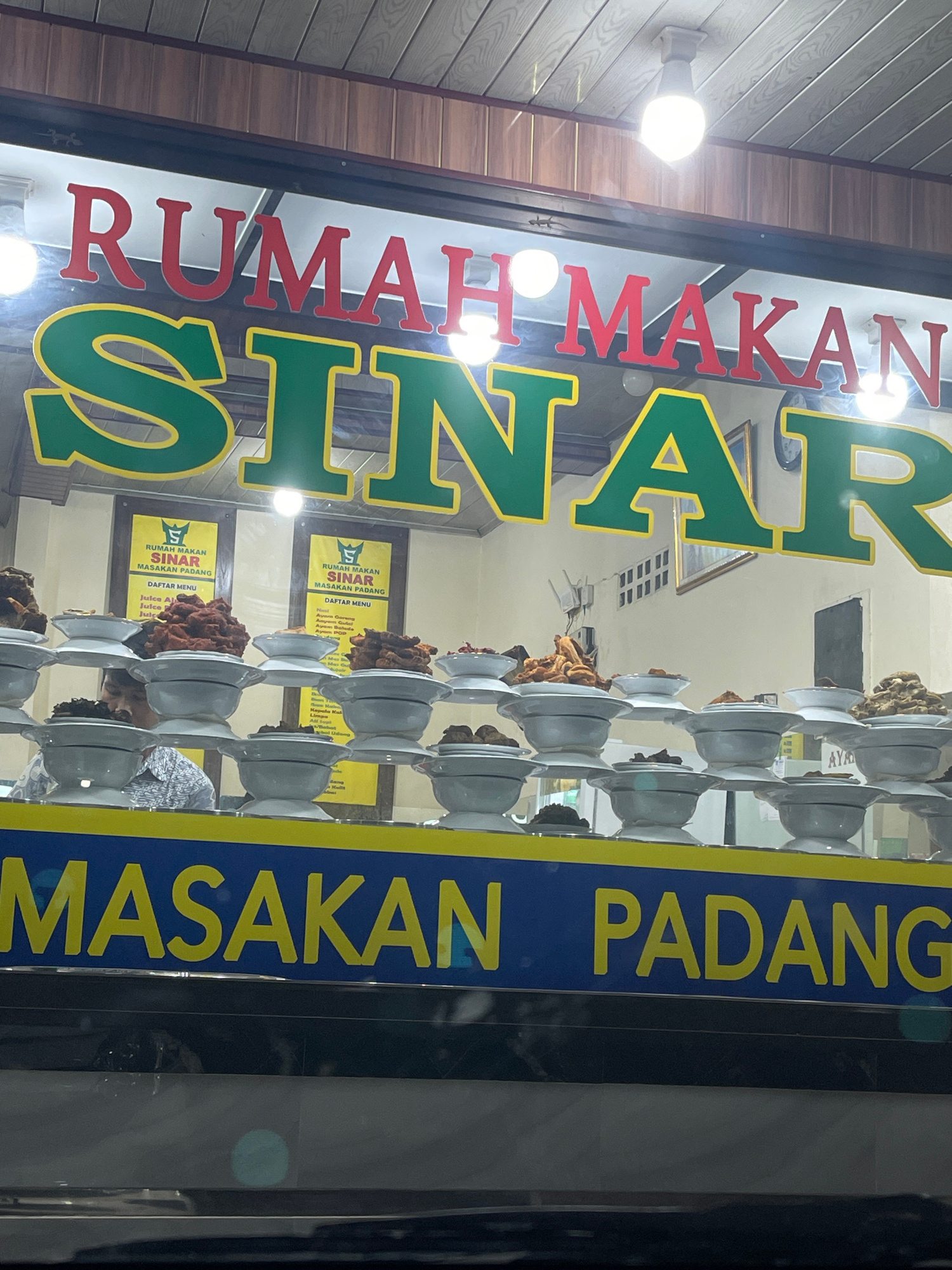 The image shows a storefront window displaying a restaurant called "RUMAH MAKAN SINAR MASAKAN PADANG." The restaurant's name is prominently displayed in large red and green letters across the top of the window. Below the name, there are stacks of white bowls filled with various food items, likely Indonesian cuisine. Two yellow signs with a logo and text are visible inside the window, one on the left and one on the right, appearing to be menus. A person, partially visible, is seen behind the counter wearing a patterned shirt. The lower part of the window features a blue banner with yellow text that reads "MASAKAN PADANG." The overall scene suggests a traditional Indonesian restaurant with a buffet-style food display.