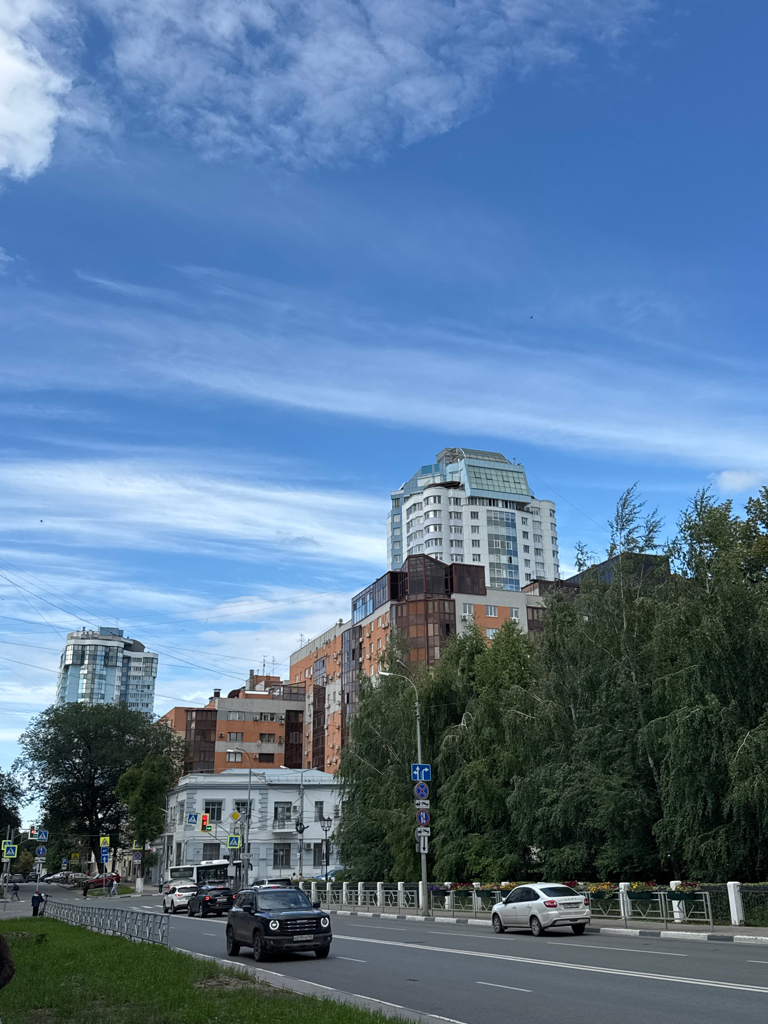 This is a medium-shot, eye-level, vertical, daytime, exterior, urban scene captured in Samara, Russia. The sky is a vibrant blue with wispy, white clouds stretching horizontally across the frame. In the foreground, a paved road runs horizontally, with a white car and a dark SUV driving towards the viewer. A pedestrian walkway with a metal fence separates the road from a green grassy area. Behind the road and walkway, a row of trees with dense green foliage lines the street. In the background, several modern multi-story buildings of varying architectural styles and colors are visible. One prominent building on the left is a tall, sleek, glass-fronted skyscraper with a unique curved design. Another building in the center is a lighter-colored high-rise with a glass roof structure. Several other residential buildings with orange, white, and grey facades are also present. Traffic lights and street signs are visible on the road. A few birds can be seen in the sky. The overall impression is a contemporary city street with residential and commercial buildings under a bright, cloudy sky.