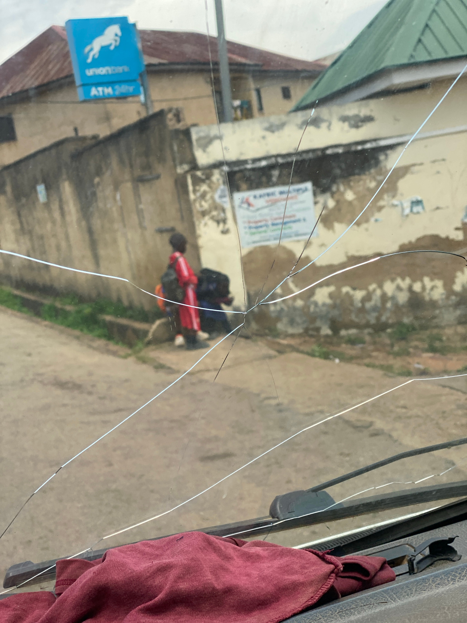 This image shows a cracked windshield from the perspective of someone inside a vehicle, looking out. The scene outside is blurry due to the cracks and the focus of the camera. In the distance, there appears to be a street with buildings and vegetation. A red motorcycle with a person on it, wearing dark clothing and a helmet, is visible in the middle ground. A blue sign with white text and a white horse logo, which reads "union bank ATM 24hr", is also visible on the left side of the frame. A maroon cloth is draped over the right side of the windshield, obscuring part of the view. The lighting suggests it is daytime, possibly a sunny day given the reflections on the glass. The overall impression is of a street scene in a developing urban or semi-urban area. The cracked windshield suggests the vehicle may have been involved in an incident.