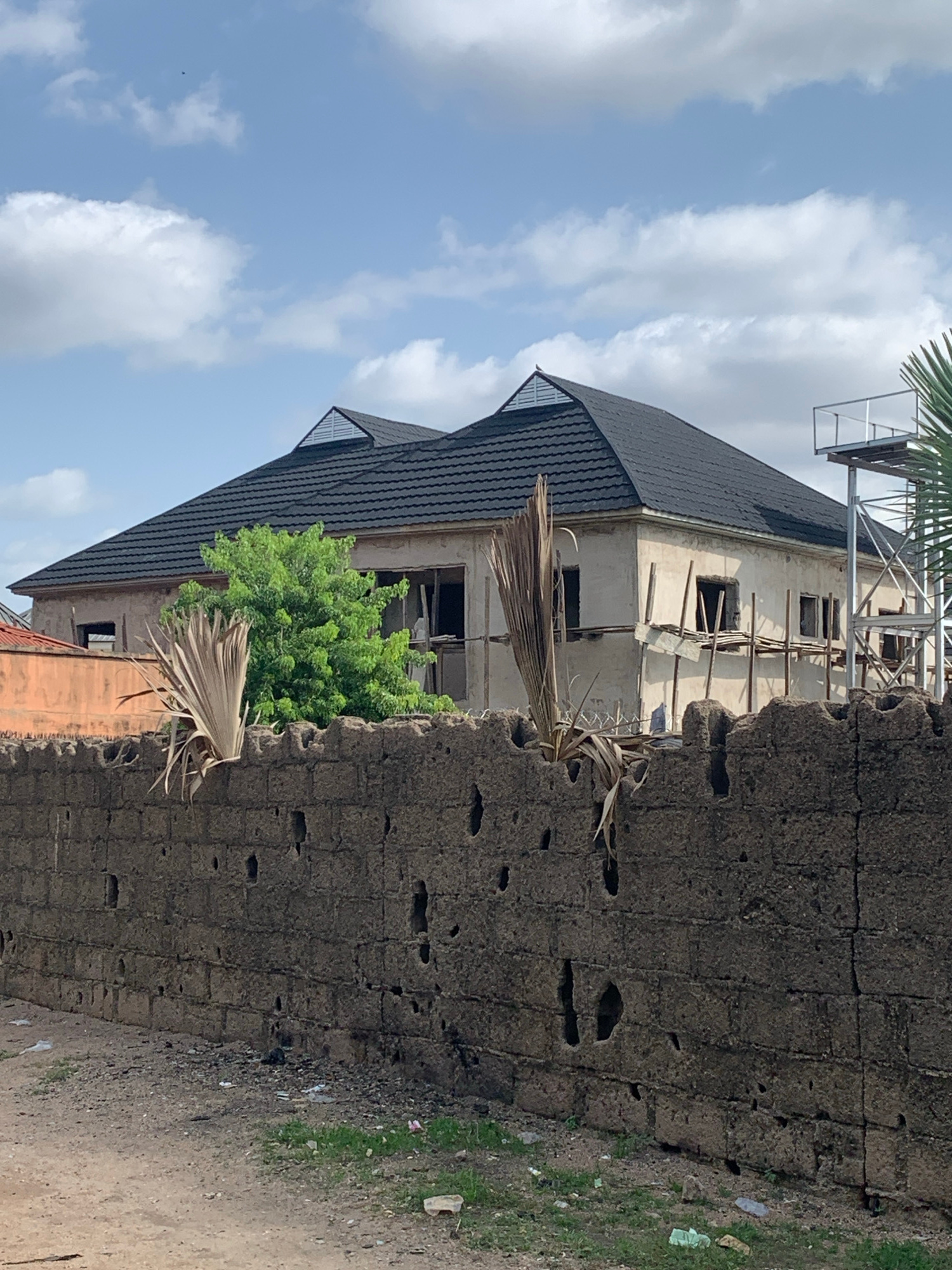 An exterior, eye-level, medium shot captures a partially constructed building with a dark, tiled roof and a rough, cement-block wall in the foreground. The building's exterior walls are a light, unfinished stucco, with visible scaffolding and openings where windows will be installed. A lush green tree is situated behind the wall and in front of the building. Two dried palm fronds extend from the top of the wall towards the building, one of them partially obscuring a window opening. To the right of the building, a metal structure, possibly a water tower or scaffolding, is visible. The sky above is blue with scattered white clouds. The ground in front of the wall is a dirt and gravel path with sparse green grass and scattered debris.