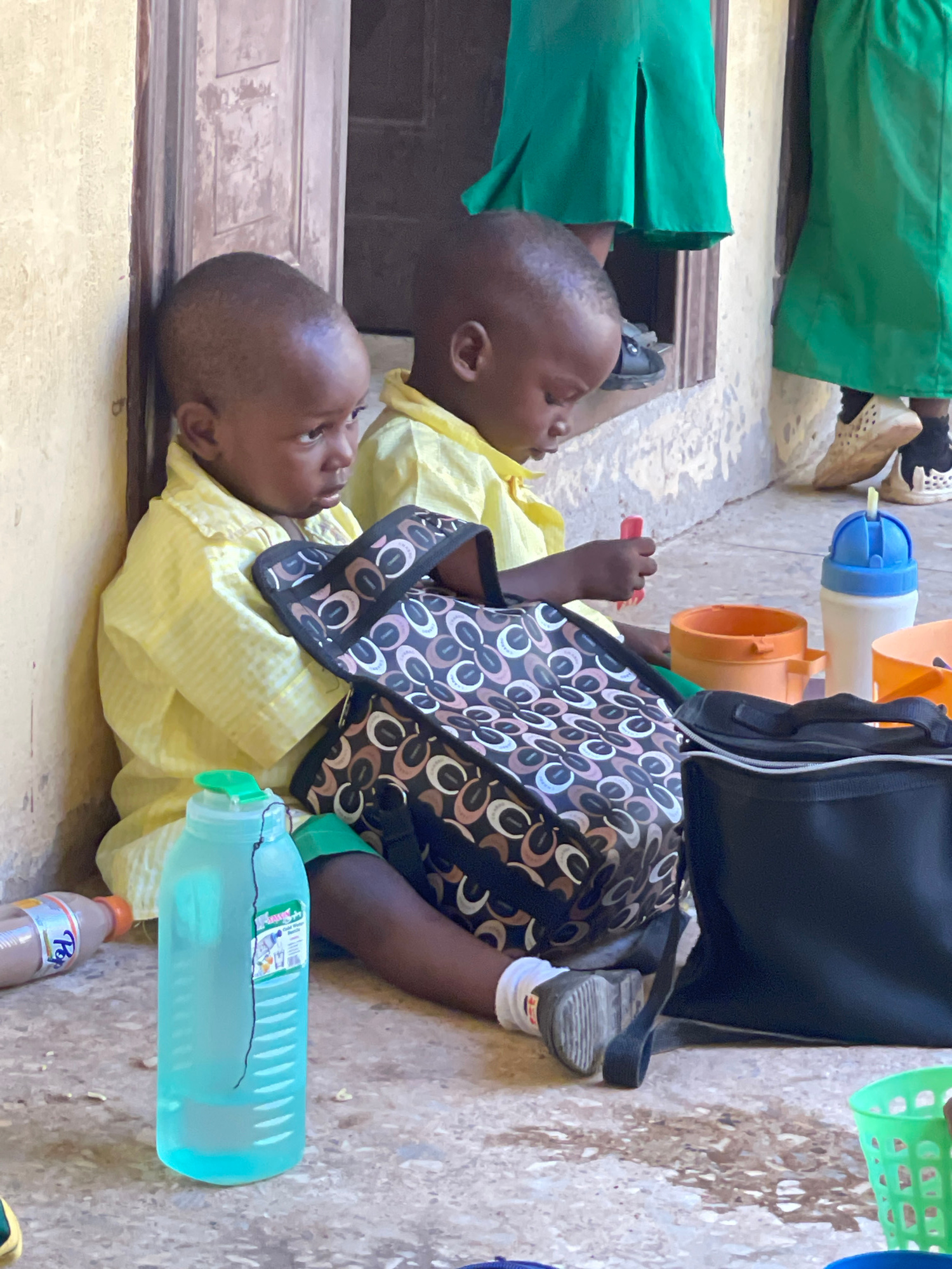 Two young children, appearing to be toddlers, are sitting on the ground in what appears to be an outdoor schoolyard or communal area in Akwanga, Nigeria. Both children are wearing yellow collared shirts, and one is also wearing green shorts. They are seated next to each other, leaning against a weathered, pale yellow wall. In front of them are various items, including a patterned bag, a large blue water bottle, an orange container, and a white and blue water bottle. The ground beneath them is paved with a speckled, light-colored material, showing some dark staining, possibly from water or dirt. In the background, other children or adults are visible, with two individuals wearing bright green skirts, suggesting a school uniform. The overall impression is one of a break time or a gathering for young students.