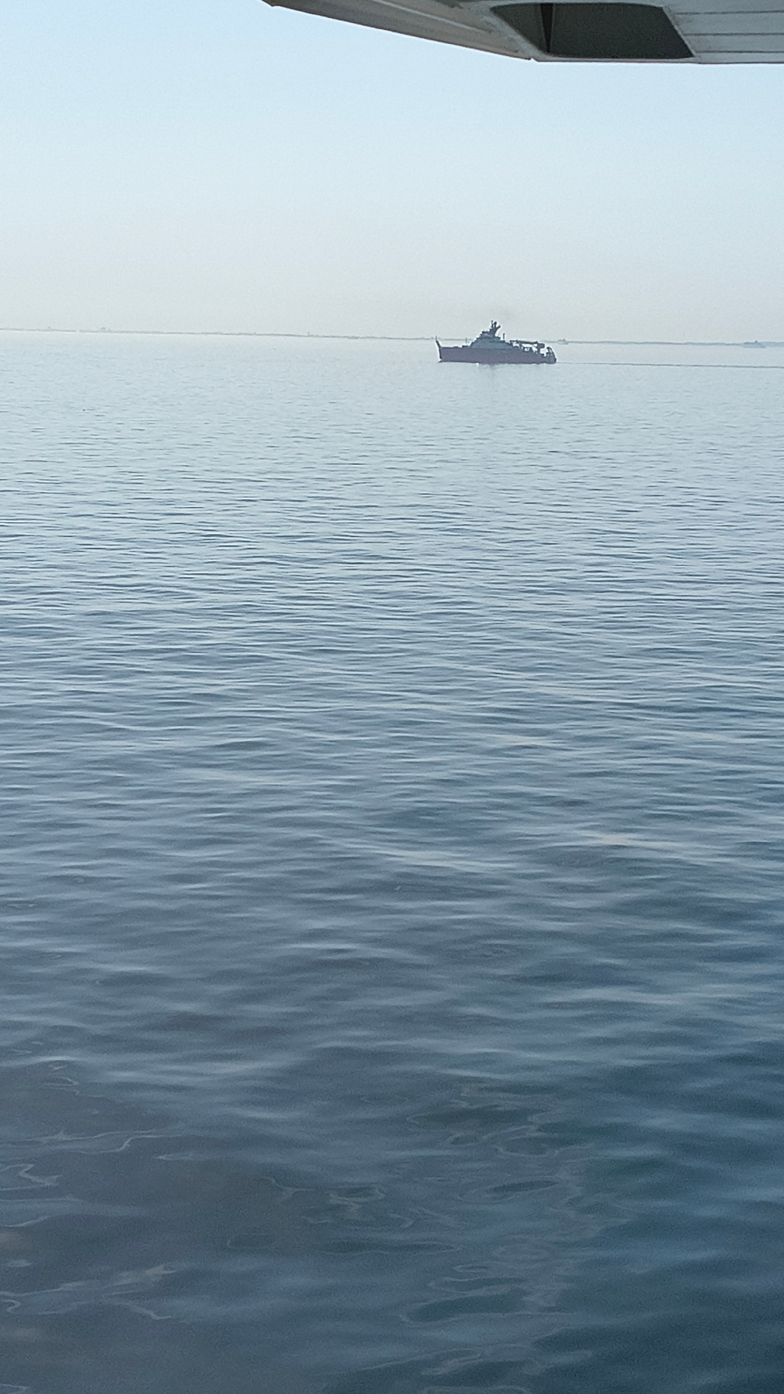 A medium-shot, eye-level, horizontal, slightly high-angle, natural light, outdoor, daytime photograph shows a large, dark-colored ship sailing on calm, blue water. The ship is located in the middle ground of the image, positioned slightly to the right of the center. It has a dark superstructure and appears to be a commercial or military vessel. The horizon line is visible in the far distance, under a pale, hazy sky. The foreground and middle ground are dominated by the surface of the water, which exhibits gentle ripples and reflections. A portion of a light-colored wing of an aircraft is visible in the upper left corner of the frame, suggesting the photograph was taken from an airplane. The scene is devoid of any human figures, and the environment is entirely natural, with no man-made structures or urban elements visible, except for the ship and the wing of the aircraft.