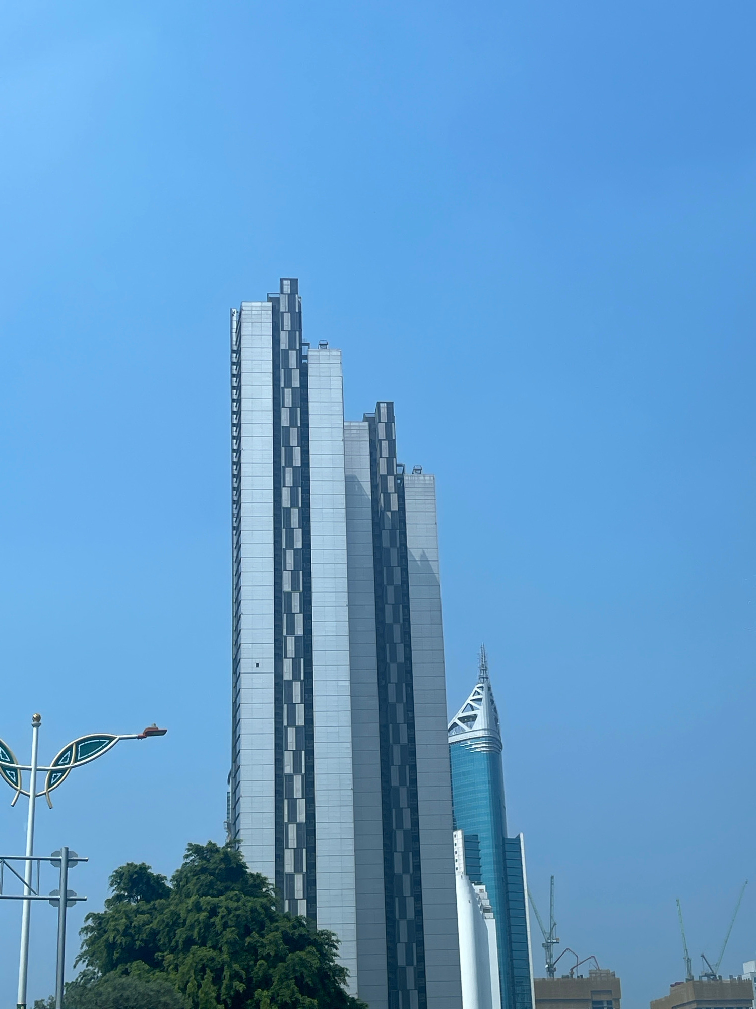 The image depicts a clear blue sky over a city skyline. In the foreground, a tall, modern skyscraper with a silver facade and dark vertical accents dominates the center-left of the frame. To the right of this building, a second, narrower skyscraper with a blue glass exterior and a pointed, white crown is visible. In the lower left quadrant, the top of a lush green tree is present, partially obscuring a decorative street lamp and traffic signals. The overall impression is one of urban development set against a bright, cloudless day. This scene is consistent with the modern architecture found in Central Jakarta, Indonesia, a district known for its high-rise buildings and commercial centers.