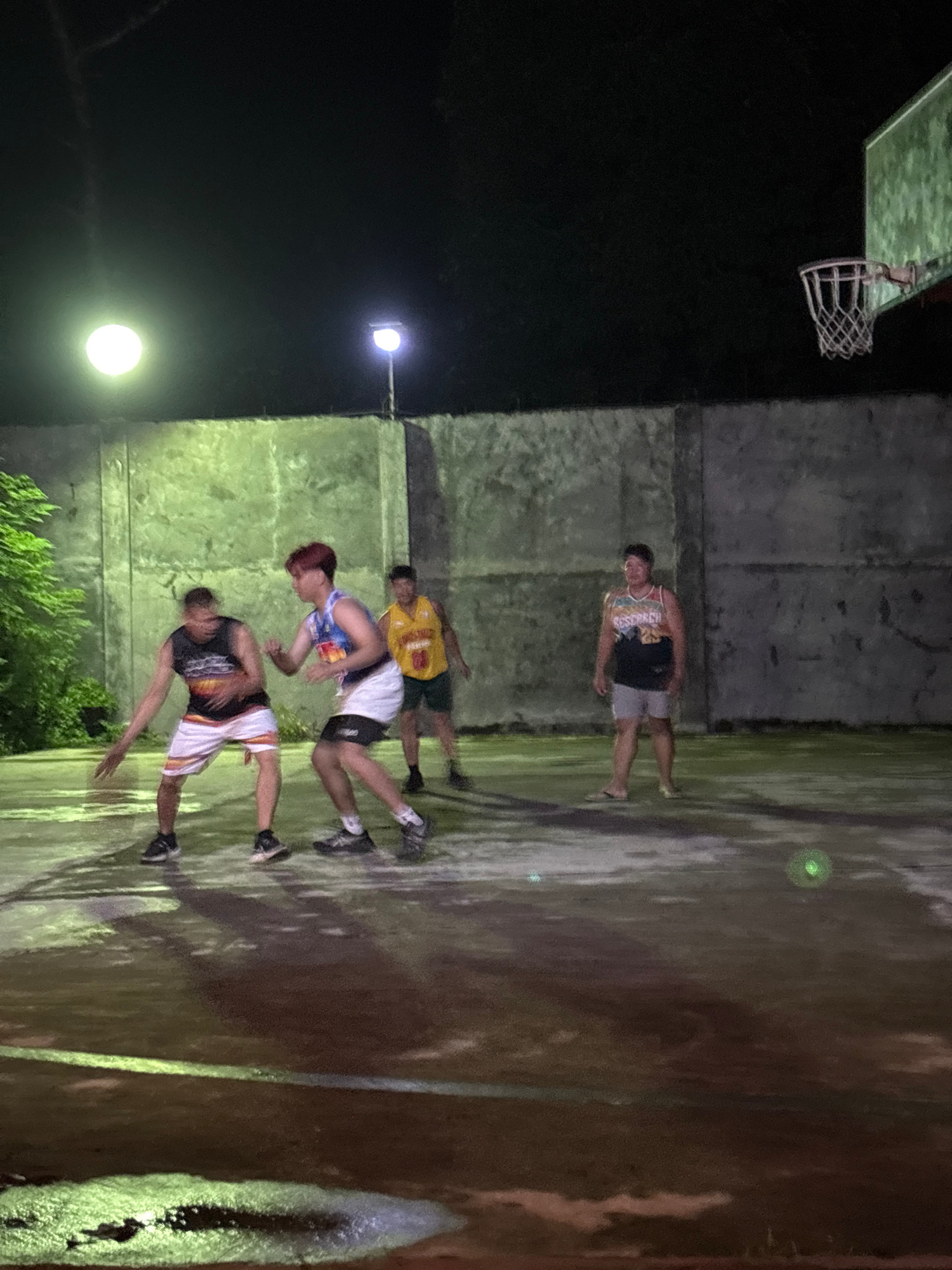 The image depicts a nighttime basketball game played outdoors in what appears to be a local court, possibly in Tagum City, Philippines. Four men are visible on the court, engaged in play. In the foreground, two players are in motion, one in a dark sleeveless shirt with colorful stripes on the shorts and the other in a white and blue sleeveless jersey. A third player, wearing a yellow sleeveless jersey, stands further back. The fourth player, in a white and colorful sleeveless top and grey shorts, is positioned on the right side of the court.
The court surface is wet, reflecting the light from overhead lamps. The background consists of a textured concrete wall and trees on the left, illuminated by outdoor lighting. A basketball hoop and backboard are visible in the upper right corner of the frame. The lighting suggests an evening or nighttime scene, with artificial lights providing illumination for the game. The overall environment is that of a casual, outdoor recreational space.