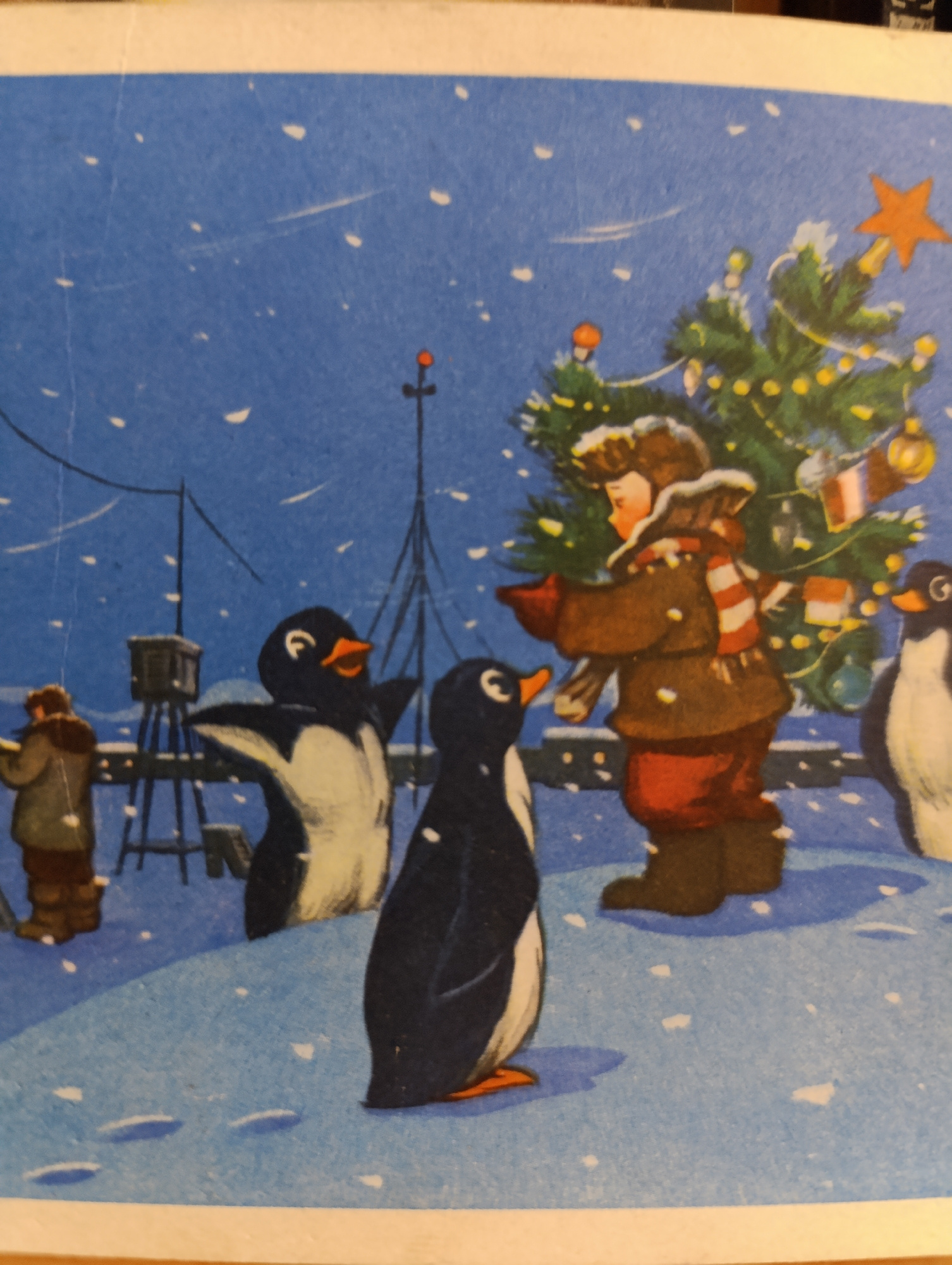 An illustration depicts a snowy, nighttime scene in an arctic or Antarctic setting. A person, appearing to be a child wearing a brown coat, red pants, and a striped scarf, is carrying a decorated Christmas tree. The tree is adorned with yellow lights and ornaments, topped with an orange star. Two penguins stand near the child, one with its wings extended. Another penguin stands further to the left, facing away from the viewer. In the background, a person in a hooded coat is visible near a structure with antennae, possibly a research station or lighthouse. The ground is covered in snow, and white flakes are falling from the dark blue sky. The scene is illuminated by the lights on the Christmas tree and a faint ambient glow.