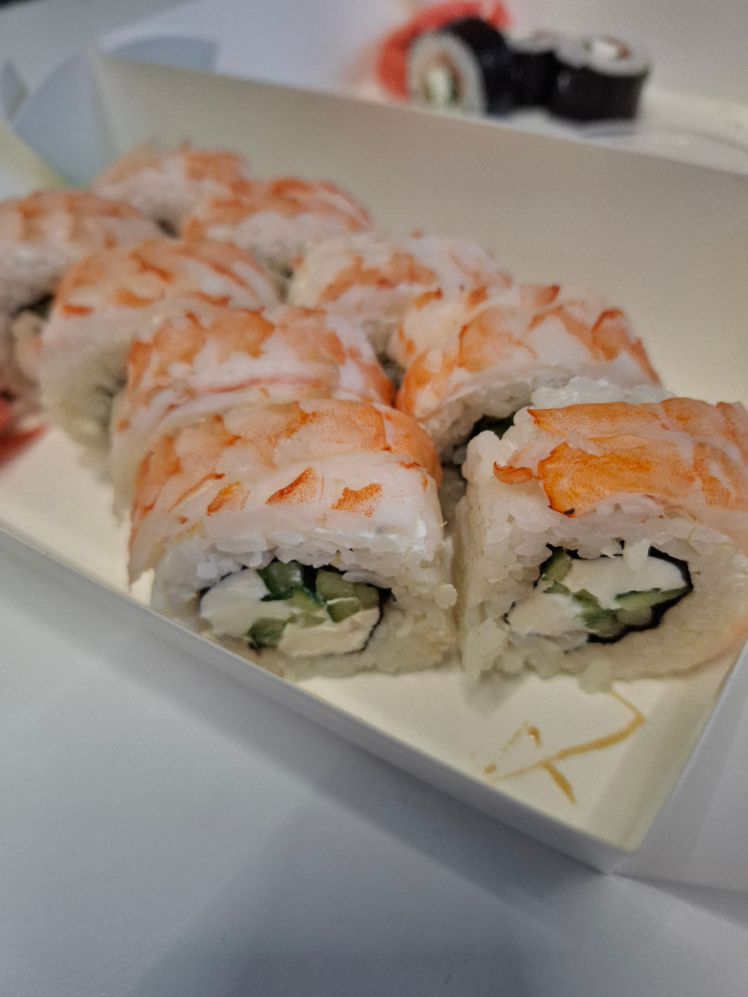 A close-up, slightly angled shot shows a white take-out container filled with sushi rolls. The rolls are made with white rice, a creamy white filling, and slices of cucumber, all wrapped in pink shrimp. Some of the shrimp pieces are translucent, while others are opaque and pinkish-orange. The sushi is arranged in a staggered pattern, with a few rolls slightly out of focus in the background, and some blurred elements suggesting other sushi items further in the background. The container itself is made of a light-colored, slightly textured material. A few faint orange markings are visible on the bottom of the container. The background is out of focus and appears to be a white surface. This image does not contain any specific geographic indicators that would allow for localization to Bratislava, Slovakia.