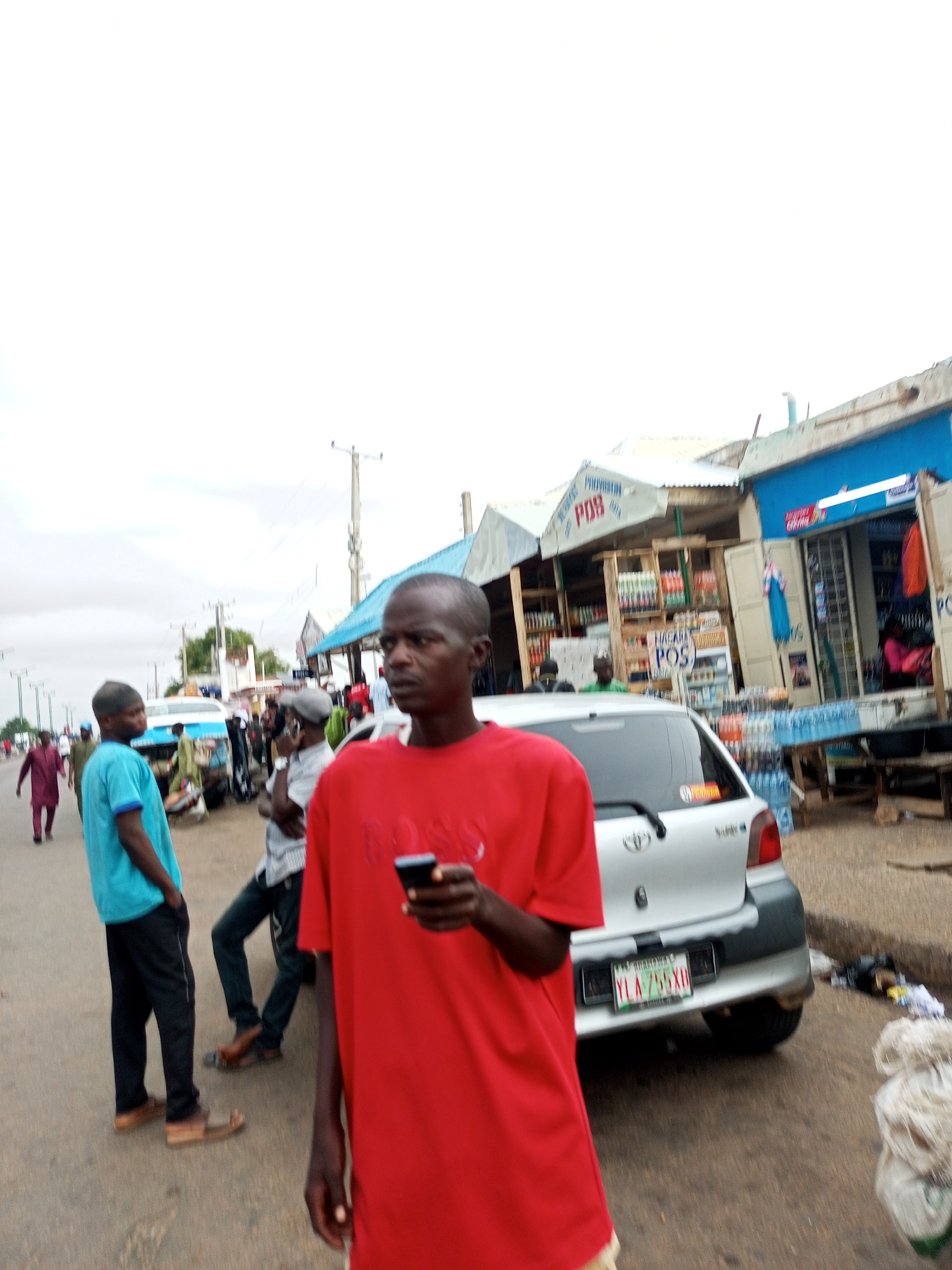 This image depicts a street scene in Gombe, Nigeria. In the foreground, a man in a red shirt is holding a smartphone. To his left, a silver car is parked, displaying a license plate that reads "YLA 755XD". Behind the car, several market stalls are visible, with various goods displayed. In the background, other individuals are walking along the street, and a few motorcycles are parked. The sky is overcast. The overall atmosphere appears to be that of a bustling marketplace or roadside vendor area.