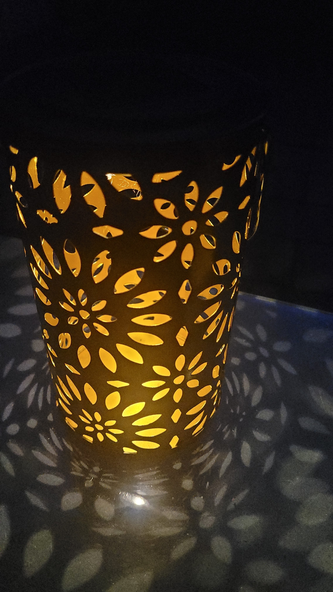 A close-up, low-angle shot shows a decorative, cylindrical lantern casting warm yellow light and intricate floral shadows onto a dark, textured surface. The lantern itself is black with a cut-out floral pattern, allowing the light from within to shine through. The shadows are cast in a symmetrical, repeating pattern of petals and blossoms, creating an atmospheric and artistic effect. The background is dark and out of focus, emphasizing the illuminated lantern and its projected patterns.