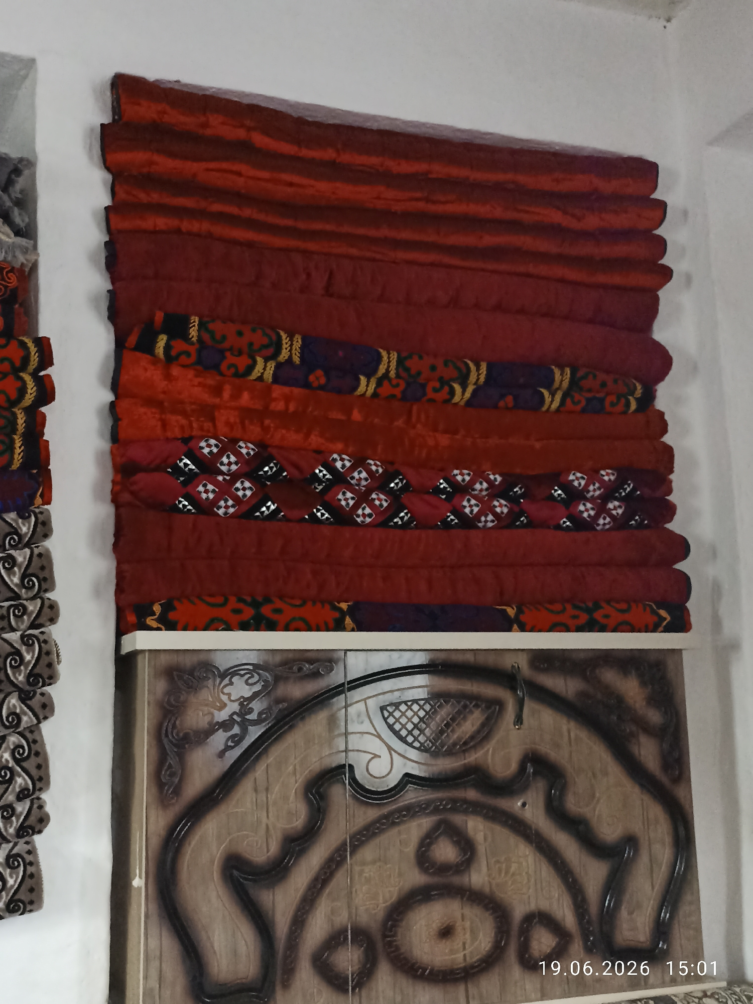 A stack of red and patterned textiles, possibly blankets or bedding, is displayed vertically against a white wall. The textiles are folded and layered, with the top portion appearing to be solid red velvet or a similar plush fabric, creating horizontal pleats. Below the solid red layers, there are bands of fabric with intricate patterns in red, black, yellow, and purple. At the very bottom of the textile stack, a band of red fabric with a repeating yellow and black geometric pattern is visible.
Beneath the textiles, a piece of furniture or a wooden surface is shown. This surface has a decorative carved or painted design that resembles an ornate medallion or crest, featuring swirling lines and geometric shapes in brown and black against a lighter wood grain background. A date and time stamp, "19.06.2026 15:01", is present in the bottom right corner of the image. To the left of the main textile stack, another narrower stack of textiles with a dark gray and white patterned design is partially visible. The overall impression is an interior scene, possibly a room or a display area, with decorative textiles and furniture.