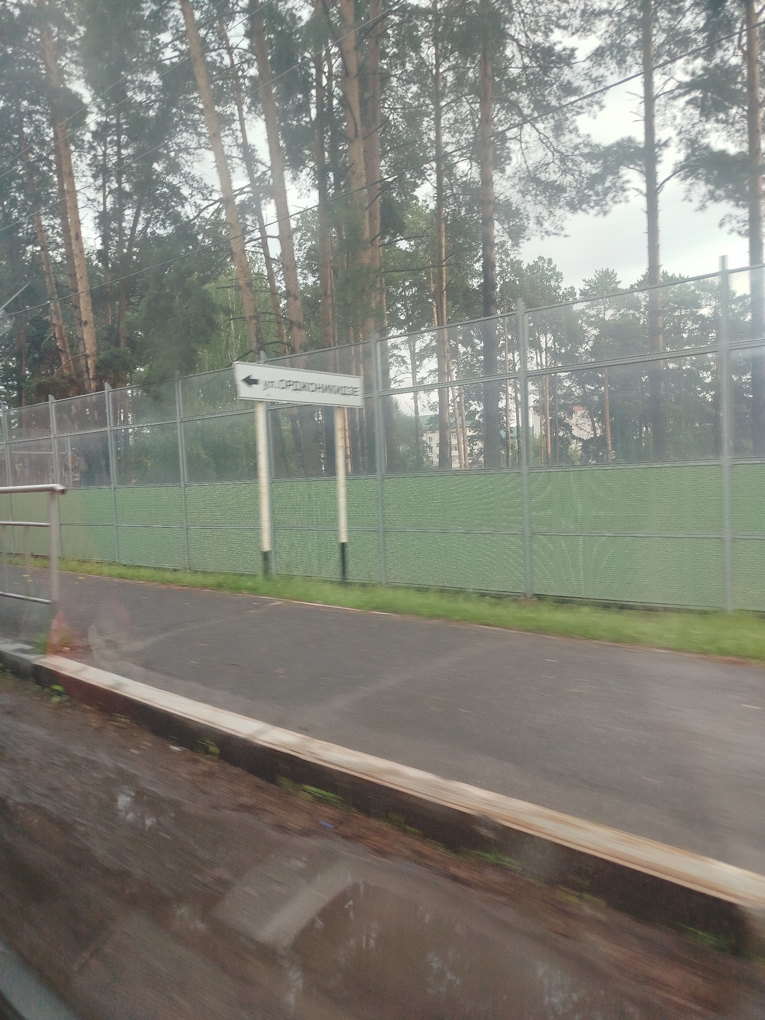 The image shows a street scene in Zavodoukovsk, Russia, with tall pine trees lining a noise barrier fence. The fence is green and extends along the side of a paved road. A signpost with a white directional arrow points to the left, indicating "ул. Орджоникидзе" (Ordzhonikidze Street). The road appears to be wet, suggesting recent rain. A metallic railing is visible on the left side of the image. The overall impression is a quiet, possibly suburban, setting on an overcast day.