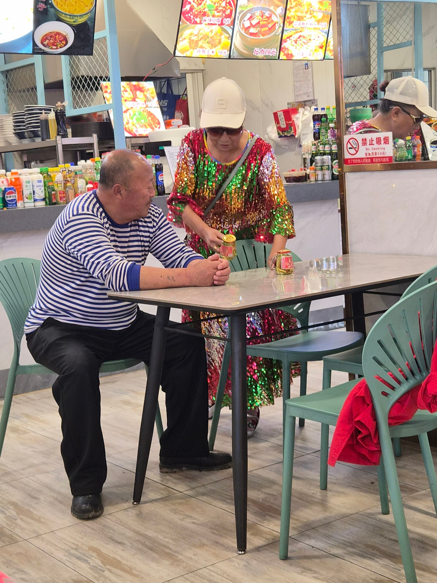 The image captures a scene inside a food establishment, likely a canteen or restaurant, in Mishan, China. A man, bald with a mustache, wearing a blue and white striped long-sleeved shirt and black pants, is seated at a table. His arms are crossed on the table, and he has a tattoo on his left forearm. Opposite him, a woman in a colorful, sequined dress and a white baseball cap is leaning forward, holding a small jar. There are other people visible in the background, including one wearing a white baseball cap and sunglasses, looking down. The environment includes tables, chairs (some green plastic, one with a red garment draped over it), and shelves stocked with various beverages and food items. A "No Smoking" sign with Chinese characters is also visible. The floor is tiled with a light wood-grain pattern. The overall impression is that of a casual dining setting within a commercial area.