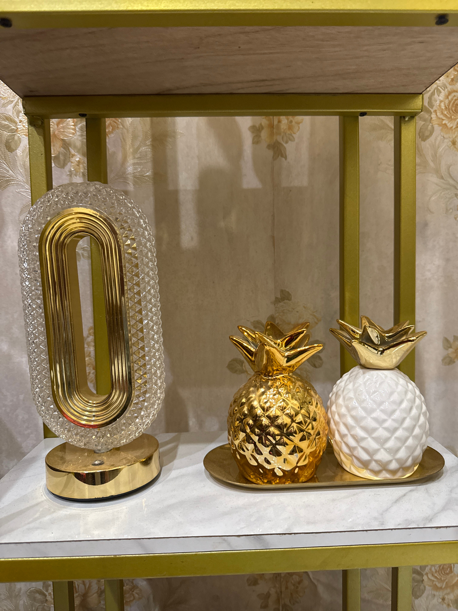 The image shows a decorative shelf with two golden pineapples, one gold and one white, and a clear, oval-shaped glass lamp with gold accents. The shelf is gold and has a wood-grain top. The background is a floral wallpaper. The scene is indoors, and the lighting is soft. No people are present, and there is no indication of the specific location within Lhokseumawe, Indonesia.