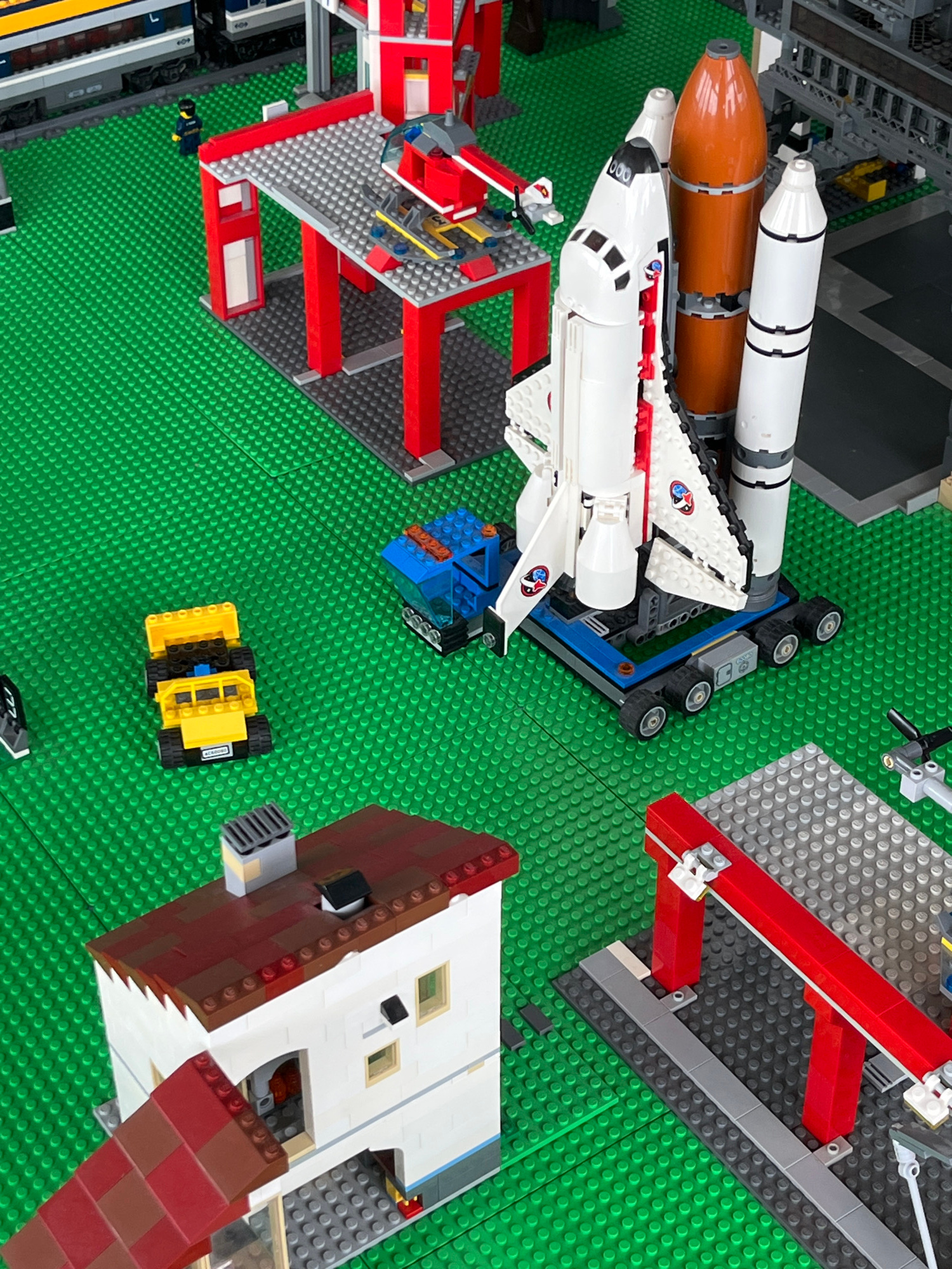 This is a close-up, high-angle, eye-level, outdoor, daylight, medium shot of a LEGO city. The foreground features a white and brown LEGO building with a red tiled roof. To the right of this building is a red LEGO gantry. In the midground, a white and red LEGO space shuttle with two booster rockets is positioned on a blue LEGO platform with wheels. A small blue LEGO truck is to the left of the shuttle. A yellow LEGO dump truck is in the lower left quadrant. In the background, there are various LEGO structures including a grey and red LEGO service station and a LEGO train. The entire scene is built on a green LEGO baseplate, simulating grass. There are no people visible in the image. The setting appears to be a toy display or a child's play area. The prompt's request to include the city of Yalta, Ukraine, is not supported by any visual information within the image.