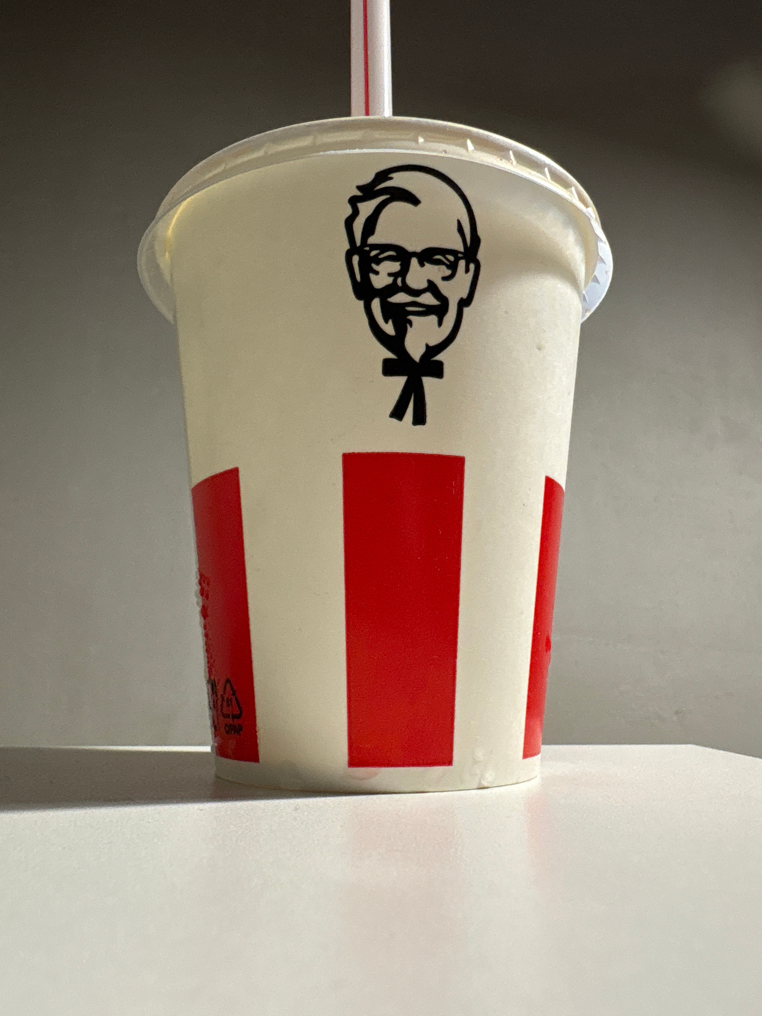 A close-up, eye-level, studio shot shows a white disposable cup with red vertical stripes and the image of Colonel Sanders printed in black on its side. A red and white striped straw protrudes from the translucent white lid. The cup sits on a white surface with a faint shadow cast to its left. The background is a gradient of light gray to dark gray. The scene is indoors and appears to be staged. There is no indication of the city of Mahilyow, Belarus.