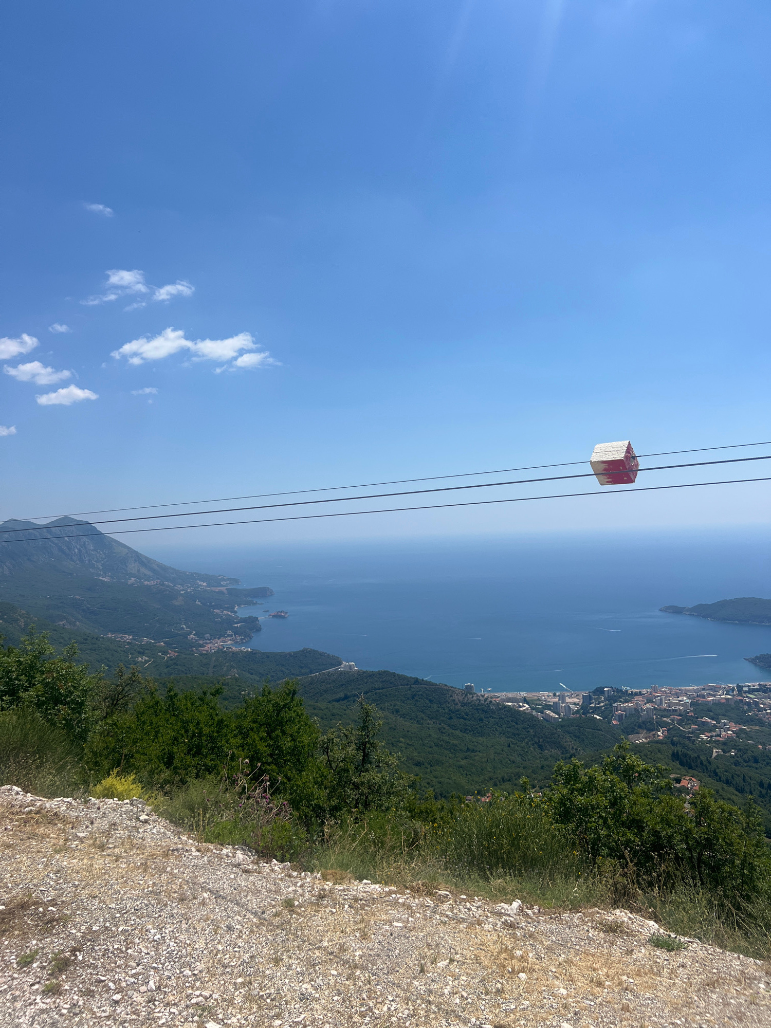 A high-angle, wide shot captures a panoramic view from a mountainside overlooking a coastal city and the Adriatic Sea under a clear blue sky. In the foreground, a gravelly embankment slopes down, bordered by green foliage and wildflowers. Three parallel wires stretch across the mid-ground, with a small, red and white octagonal object suspended from them. Beyond the wires, the lush green slopes of mountains descend towards the city, which is nestled along the coastline. Buildings, some with red roofs, are visible interspersed with greenery. The deep blue sea stretches to the horizon, dotted with a few white boats. The hazy silhouettes of distant mountains frame the scene. The image appears to be taken from the vicinity of Brajici, Montenegro, possibly from the cable car route to the Lovcen National Park. There are no people visible in the image.