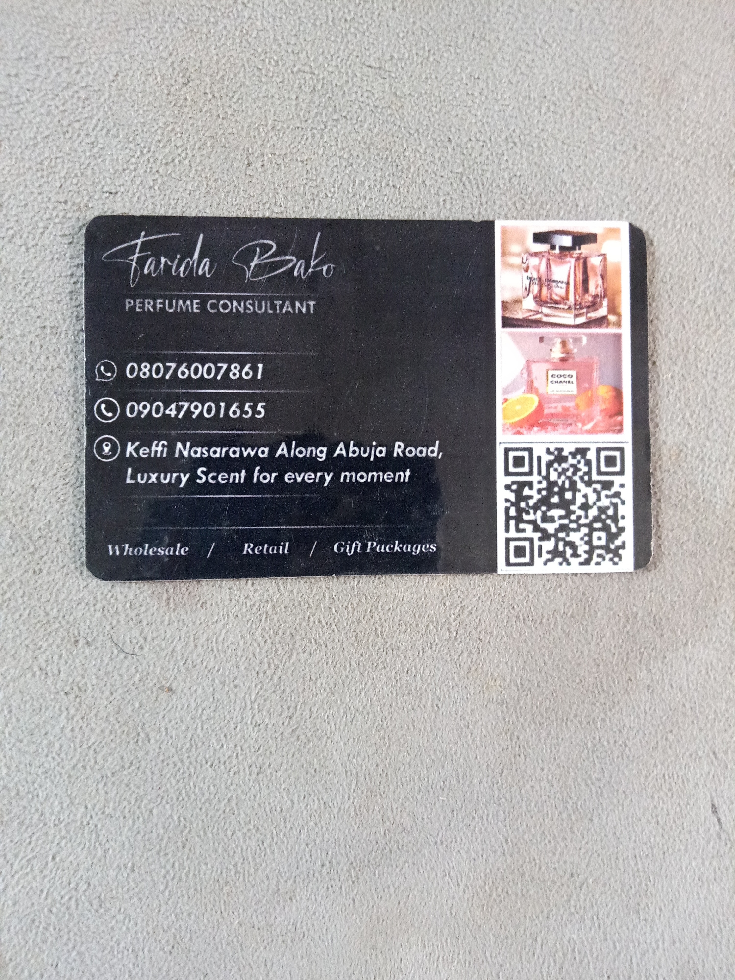 A black business card is displayed on a textured, light gray surface, possibly concrete. The card features white text and a QR code. The text on the card reads "Farida Bako PERFUME CONSULTANT" followed by a phone number "08076007861" and another phone number "09047901655." Below the phone numbers, an address is listed as "Keffi Nasarawa Along Abuja Road, Luxury Scent for every moment." At the bottom of the card, the words "Wholesale / Retail / Gift Packages" are visible. To the right of the text, there are two small images of perfume bottles and a QR code. The business card appears to be the main subject of the image, with the background providing context for its presentation.