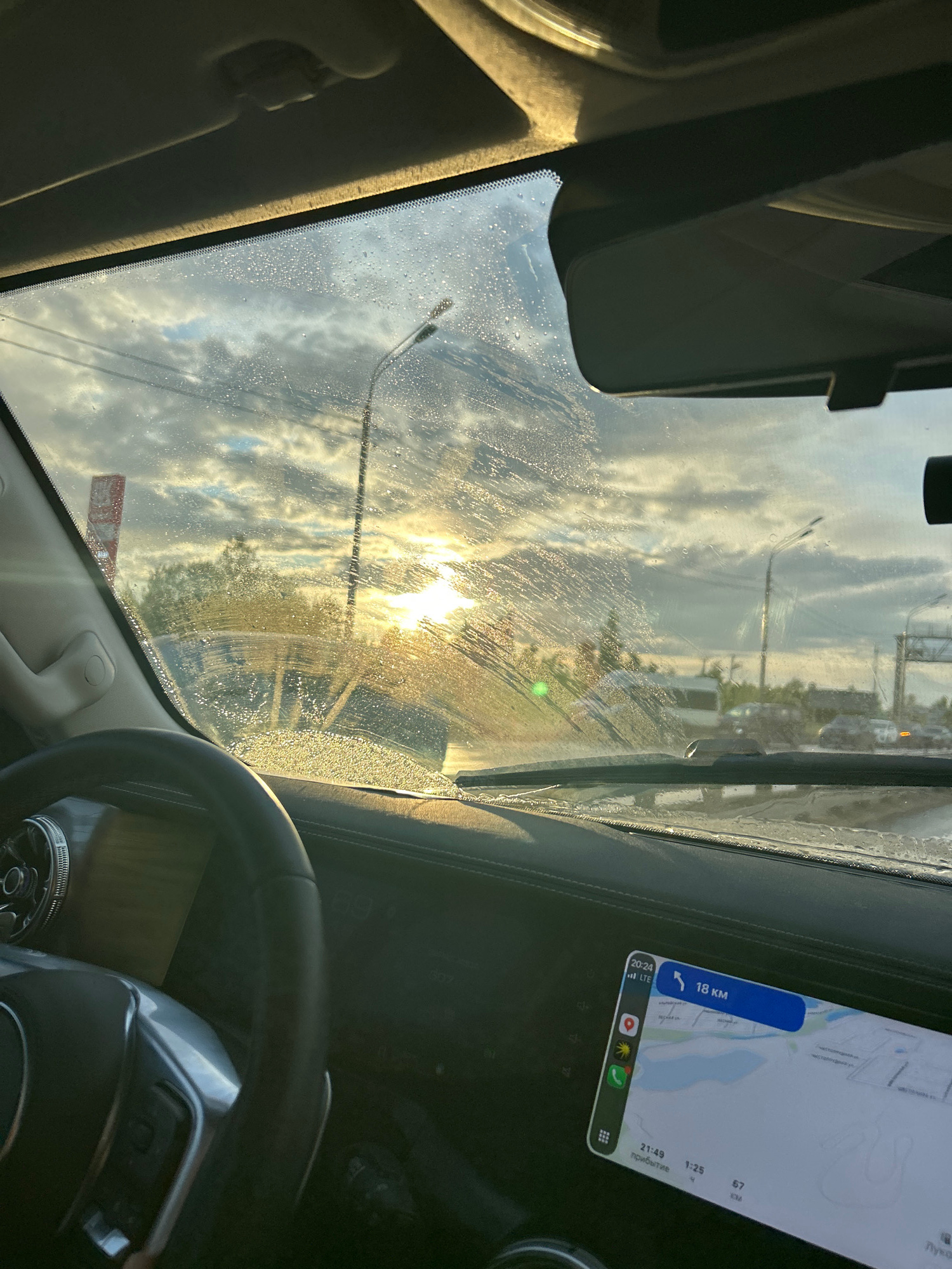 The image depicts the view from inside a car, looking through the windshield. The sun is shining brightly, creating a glare on the wet windshield, which has streaks and water droplets. The sky is cloudy with patches of blue. In the distance, there are streetlights and trees, suggesting an outdoor, possibly urban or suburban, setting. A car's steering wheel is visible in the foreground on the right side. A smartphone mounted on the dashboard displays a navigation app showing a map, with the text "18 KM" indicating distance and "20:24" showing the time. Other visible text on the screen includes "LTE", "1:25", "67 KM", and what appears to be Cyrillic text reading "прибытие" (arrival). The overall impression is of a car journey during the late afternoon or early evening, with the weather being damp or recently rained. The location is identified as посёлок опытного хозяйства «Ермолино», Russia.