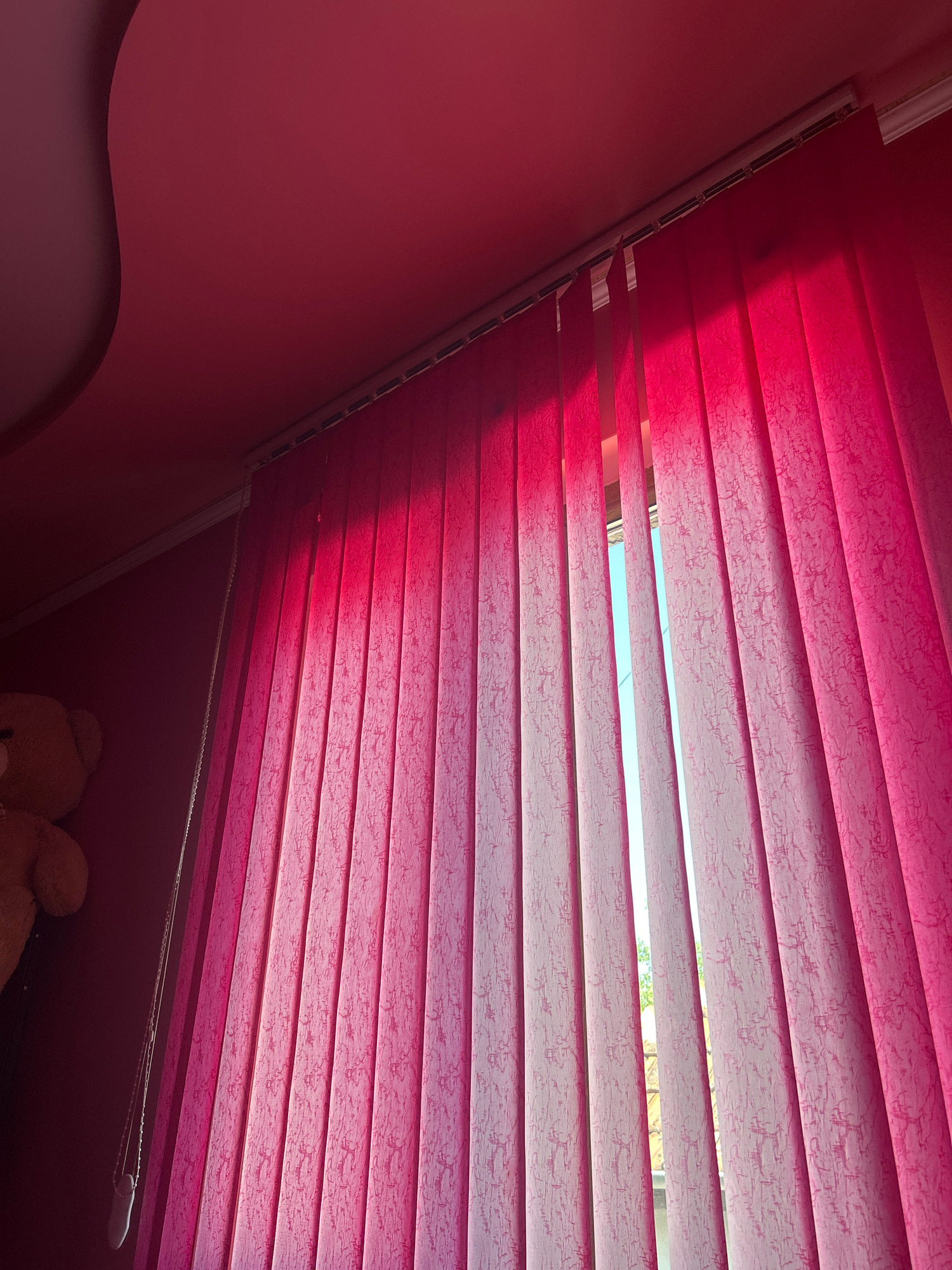 A teddy bear is partially visible on the left side of the image, positioned near a wall. In the foreground, vertical blinds, predominantly pink with some lighter textured patterns, cover a window. The blinds are angled, allowing a view of a bright sky with some hints of greenery through the gaps. The ceiling is visible at the top of the image, with a curved architectural feature on the left. The overall lighting suggests a bright, daytime scene, possibly indoors. The location is identified as Knezha, Bulgaria.