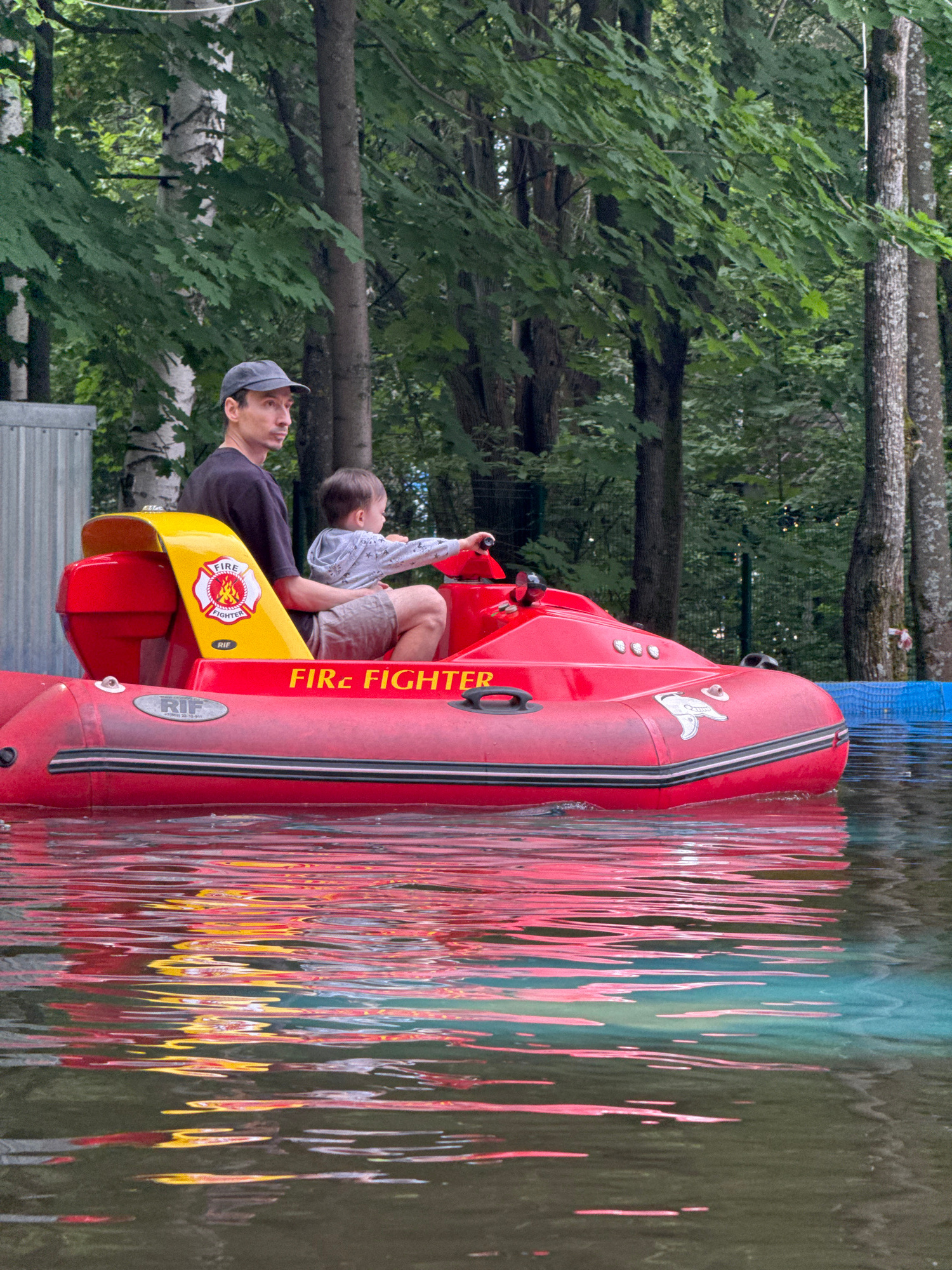 This is a vertically oriented, medium-shot photograph taken outdoors. The primary subjects are two people: an adult male wearing a dark t-shirt and a baseball cap, and a young child. They are seated in a bright red inflatable boat, designed to resemble a fire truck, which is floating on water. The boat has yellow accents and features logos with the text "FIRE FIGHTER" and "RIF". A white bird emblem is also visible on the side of the boat.
The scene is set against a backdrop of lush green trees and foliage, suggesting a park or natural environment. The water's surface is rippled, reflecting the red and yellow colors of the boat and creating a vibrant, abstract pattern. The reflection also shows hints of a blue structure in the background, possibly part of a ride or attraction. The lighting indicates it is daytime, with a slightly overcast or diffused light, as there are no harsh shadows. The overall mood appears to be relaxed and recreational, likely a family outing at an amusement park or water attraction.
Visible text: "FIRE FIGHTER" and "RIF". The location context is Izhevsk, Russia.