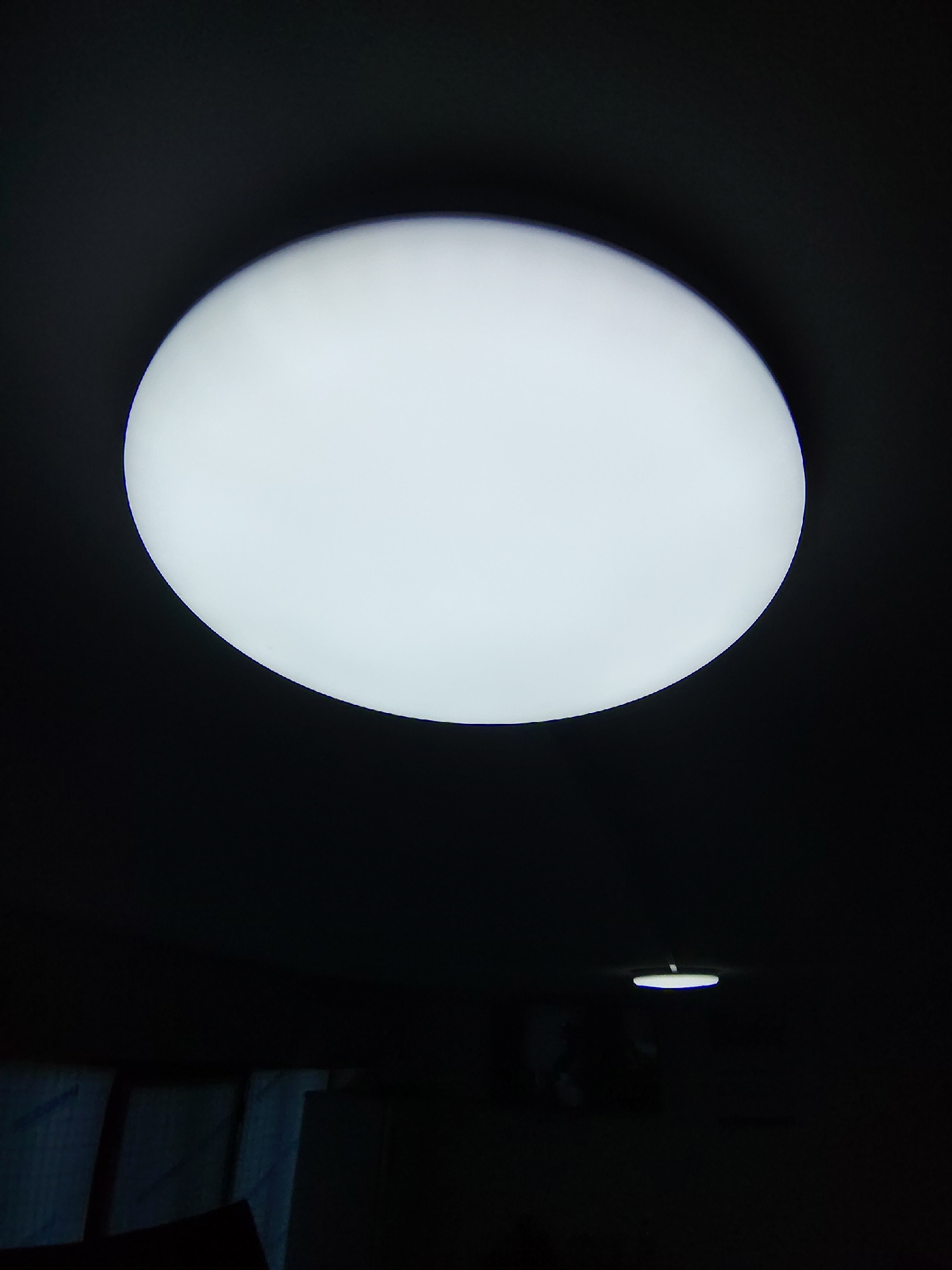A bright, round light fixture dominates the upper half of the image, casting a soft white glow against a dark background. The light appears to be a ceiling-mounted fixture. In the lower portion of the image, a much smaller, dimly lit light fixture with a blueish hue is visible in the distance. Below that, the dark shapes of what appear to be furniture or possibly curtains can be discerned. The overall scene suggests an indoor environment at night or in low light conditions. The image does not contain any specific details that would allow for identification of its location within the city of Manguzar, Uzbekistan.