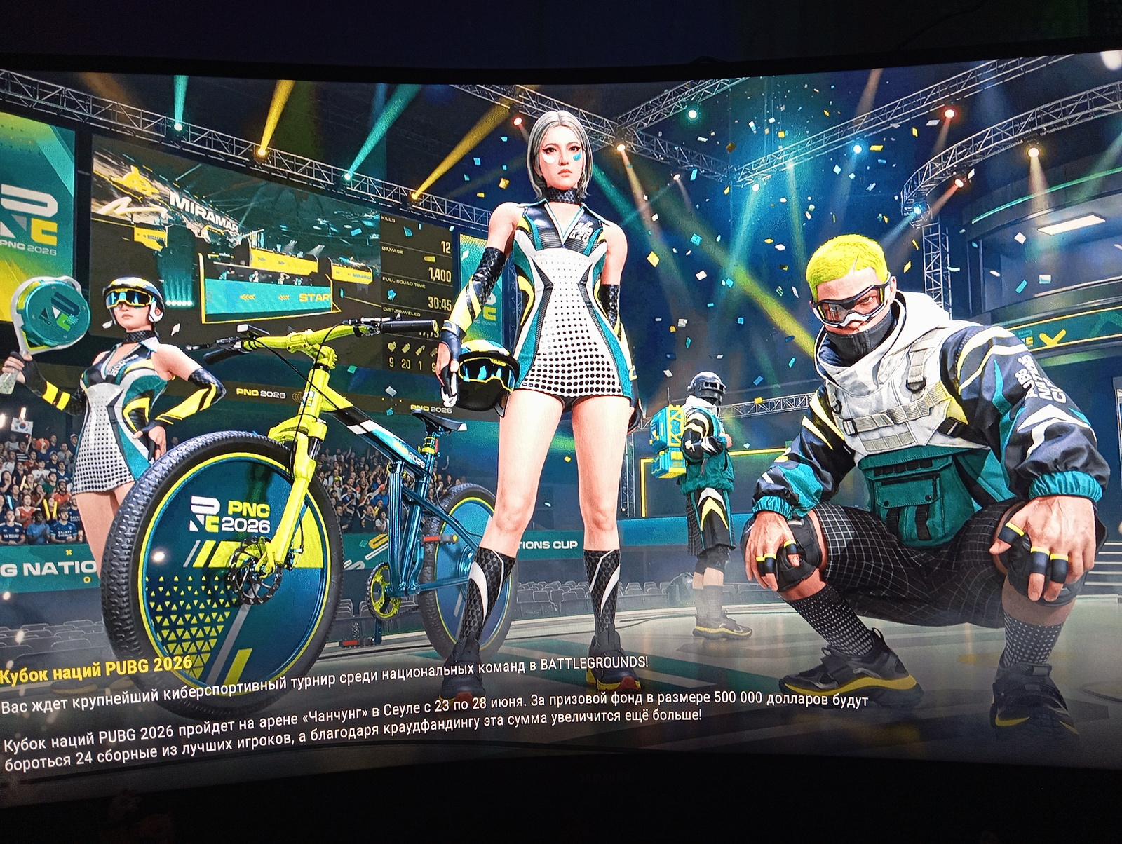 The image displays a scene from a video game, likely a loading screen or menu for "PUBG Nations Cup 2026". In the foreground, three individuals are featured. On the left, a female character with blonde hair, wearing a futuristic outfit with teal, white, and black accents, holds a green and yellow circular object. To her right is a bright yellow bicycle with a distinctive wheel design. In the center, the same female character is shown from the waist down, wearing a similar outfit and black boots. To the right of the female character, a male character with bright yellow hair, wearing a white hooded jacket over a teal and black suit with yellow accents, is crouching. He is wearing goggles and black fingerless gloves. In the background, a large stadium-like arena is depicted with multiple levels of seating filled with spectators. Bright spotlights illuminate the scene, and confetti is falling. On a large screen in the background, the word "MIRAMAR" is visible, suggesting a desert map in the game. The overall aesthetic is vibrant and energetic, indicative of a competitive esports event. Text in Russian is visible at the bottom of the screen, indicating that the "PUBG Nations Cup 2026" will take place in Seoul, South Korea, from June 23rd to June 28th, with a prize pool of 500,000 US dollars, which will increase further due to crowdfunding. There is no indication of Sanremo, Italy, in the image.