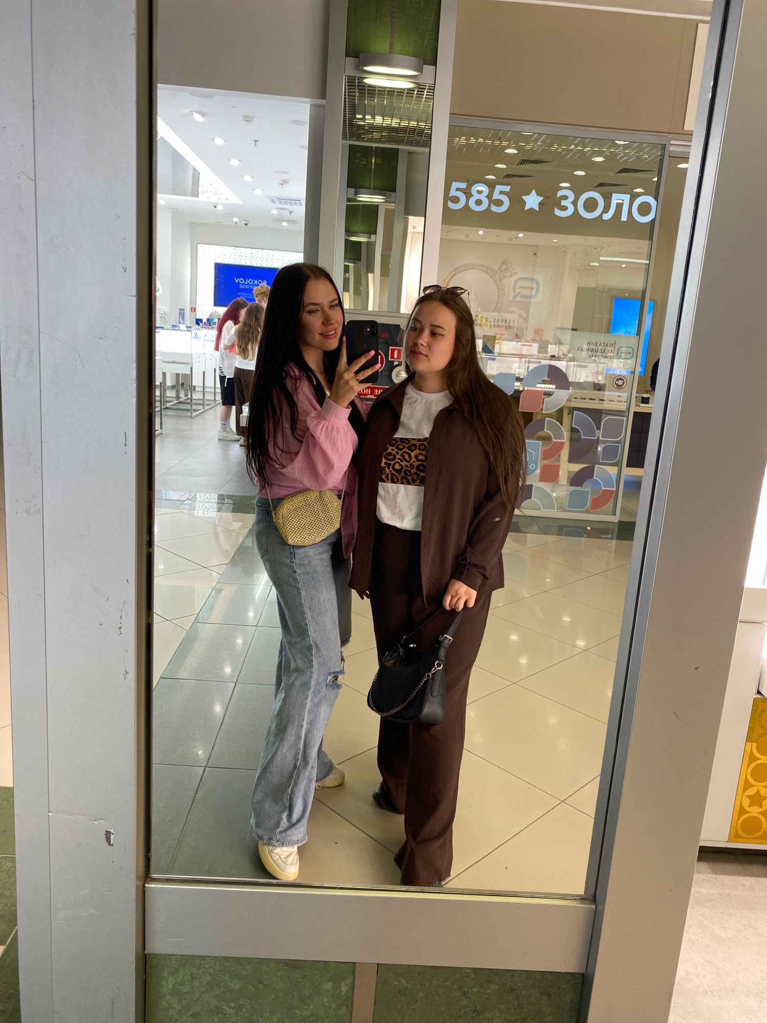 Two young women are taking a mirror selfie inside a brightly lit retail store. The woman on the left has long dark hair and is wearing a pink shirt and ripped blue jeans. She is holding a black smartphone and has a small straw purse slung across her body. The woman on the right has long brown hair and is wearing a brown pantsuit over a white t-shirt with a leopard print panel. She is carrying a black handbag with a gold chain strap. In the background, a large sign reads "585 ЗОЛОТО", which translates to "585 Gold". This suggests the location is likely a jewelry store. The floor is tiled with a checkered pattern. The store appears to be in a modern shopping mall in Perm, Russia, as indicated by the presence of the "585 ЗОЛОТО" chain, which is a prominent jewelry retailer in Russia, and specifically the presence of this chain in Russia makes this an accurate assumption.