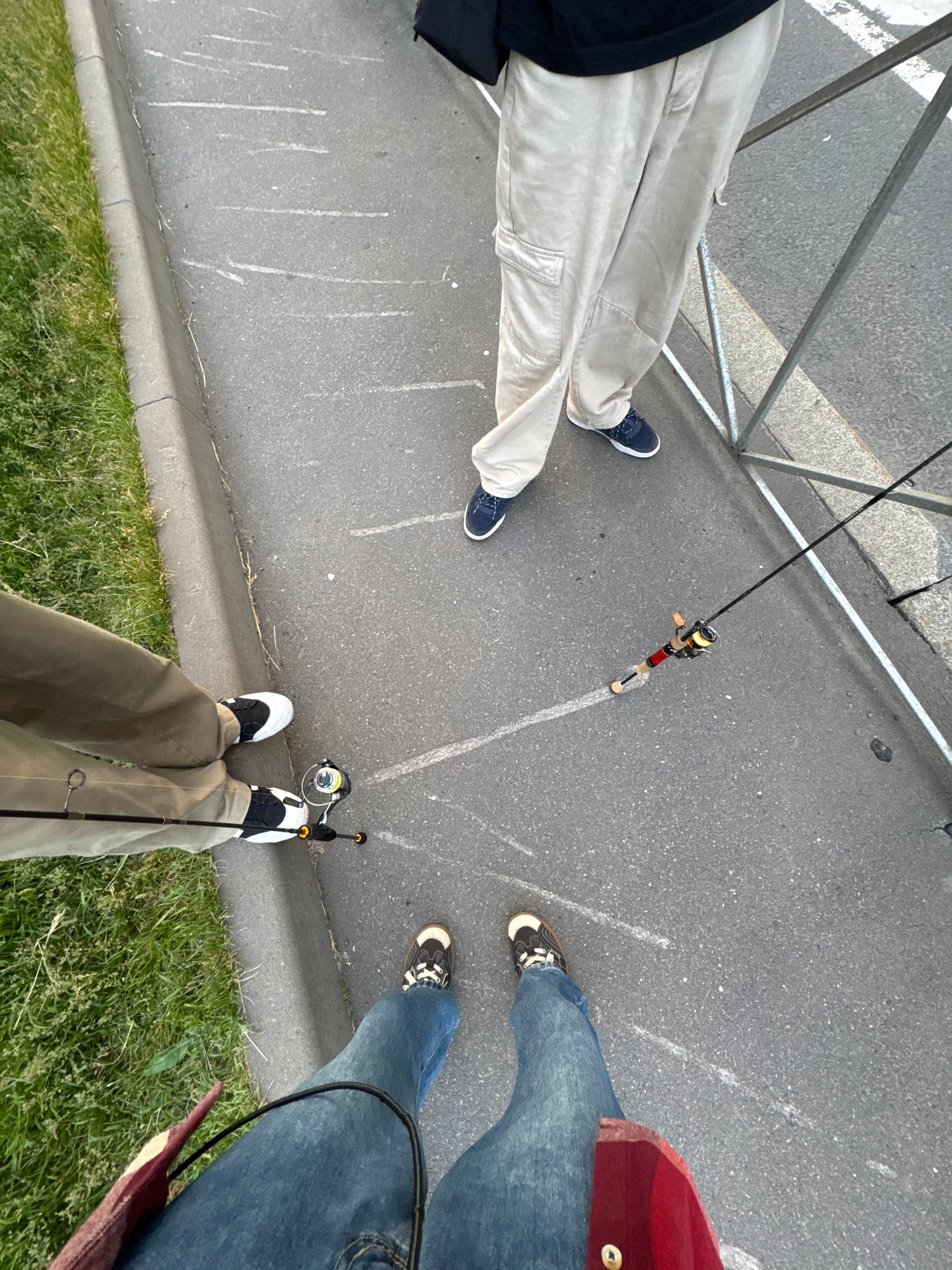 Three individuals are visible from the knees down, standing on a paved surface with a grassy area to the left. One person wears light-colored cargo pants and dark blue sneakers. Another wears khaki pants and white and black sneakers. The third person wears blue jeans and dark-colored sneakers with brown accents. Two fishing rods are present; one is red and gold, and the other is black with a silver reel. The scene appears to be a sidewalk or walkway, possibly near a park or waterfront, given the presence of grass and pavement. There are no discernible landmarks that definitively place the location in Saint Petersburg, Russia, although the overcast sky and general urban aesthetic could be consistent with the city.