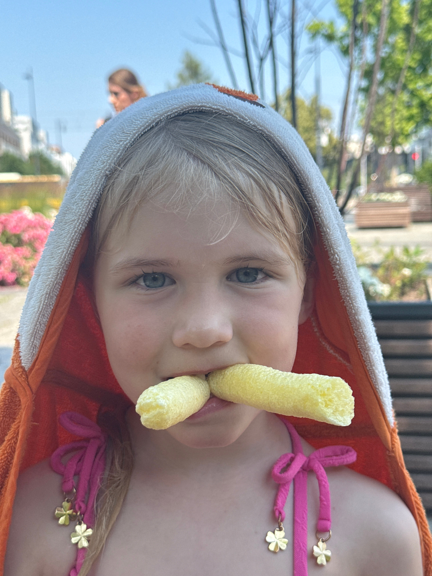 The image shows a close-up, tilted shot of a young girl wearing an orange hooded towel with white trim. She has blonde hair and blue eyes, and she is holding two yellow corn puff snacks in her mouth, with one partially obscuring her lips. She is wearing a pink bikini top with decorative gold clover charms. The background is blurred, showing greenery and a wooden structure, suggesting an outdoor setting. The lighting indicates it is daytime, likely sunny. The girl's expression is playful and engaging. In the background, another person's shoulder and head are visible. The setting appears to be a public space, possibly a pool area or garden, in Warsaw, Poland. There is no visible text in the image.