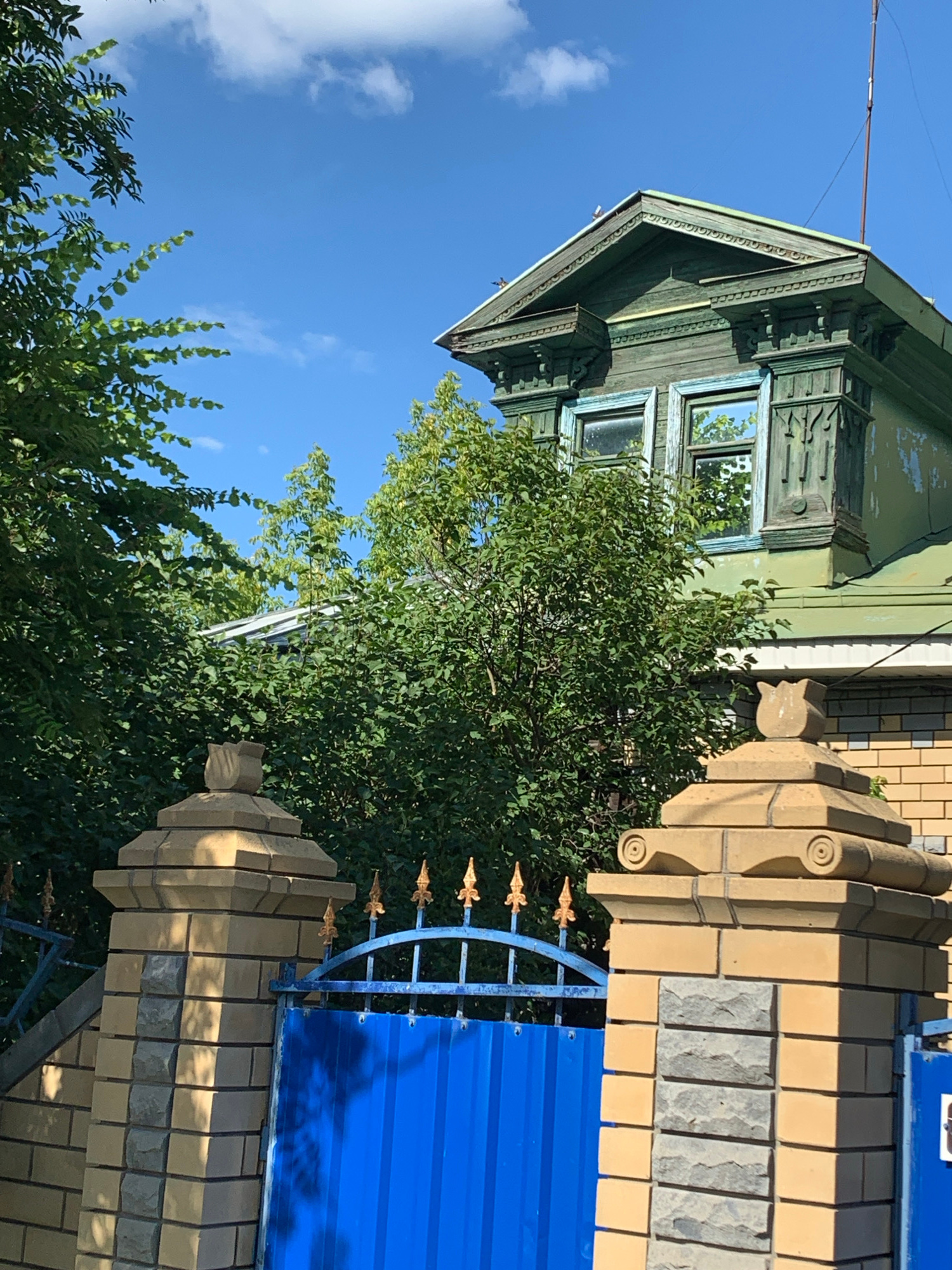 The image captures an exterior view of a traditional Russian wooden house, painted green and showing signs of age, with ornate trim around the windows and eaves. The house is situated next to a fence made of light-colored bricks and topped with a bright blue metal sheet. Decorative elements are visible on the brick pillars and a golden metal gate. Lush green trees and foliage surround the property, suggesting a natural or garden setting. The sky is a clear, bright blue with a few scattered white clouds, indicating a sunny day, likely during daylight hours. The composition and subject matter suggest a residential setting, possibly in a rural or suburban area of Pavlovo, Russia. There is no visible text in the image.