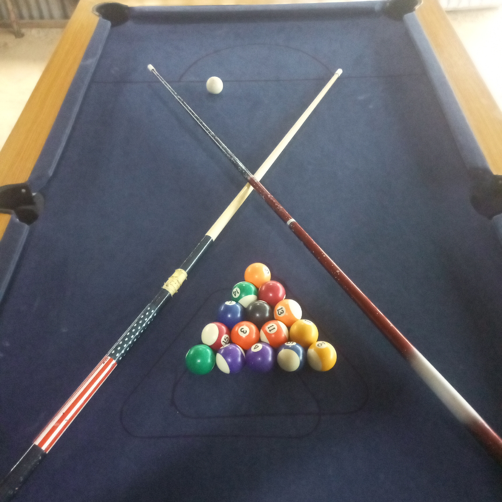The image shows a pool table with billiard balls arranged in a triangle formation and two pool cues resting diagonally across the table. One cue has a design resembling the American flag, with red and white stripes and a blue section with white stars. The other cue is a plain reddish-brown color with a white section near the tip. A white cue ball is positioned on the table near the top of the triangle of balls. The billiard balls are numbered from 1 to 15, with various colors, including solid colors and striped balls. The pool table has a dark blue felt surface and a wooden frame. The setting appears to be indoors, possibly in a recreational area or home. The lighting suggests it could be daytime. There is no visible text other than the numbers on the billiard balls. The scene depicts a static setup, with no discernible activity or event taking place beyond the preparation for or aftermath of a game of pool. The image is taken from a slightly elevated perspective, looking down onto the table.