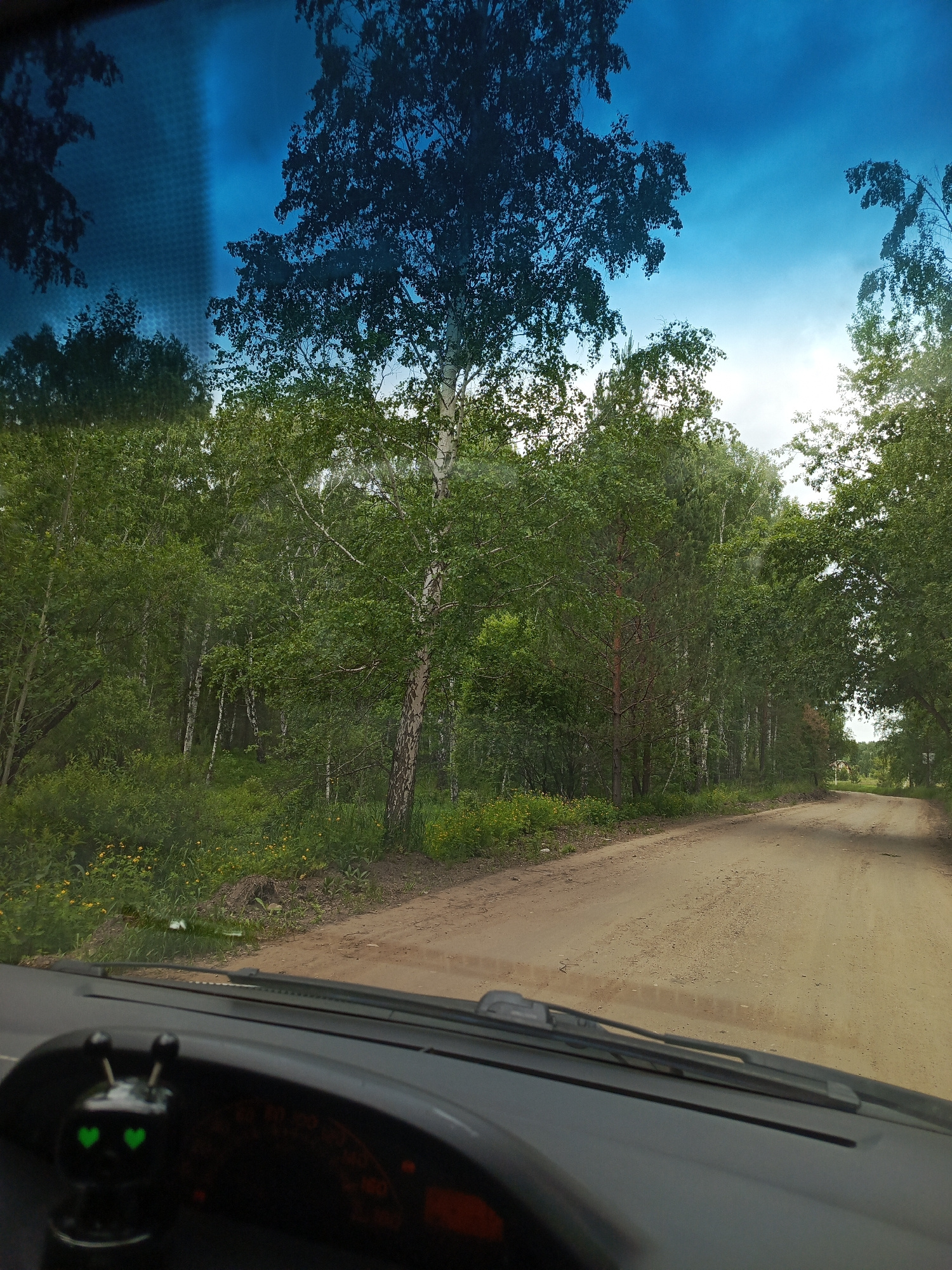 The image is a first-person perspective shot from inside a vehicle, looking through the windshield. A dirt road curves to the right through a dense forest. The trees are predominantly deciduous with green foliage, and some birch trees are visible with their characteristic white bark. The sky is a bright blue with some white clouds. The dashboard of the vehicle is visible in the foreground, with a small black ornament with green heart-shaped eyes positioned on the left side. The location is identified as Meget, Russia.