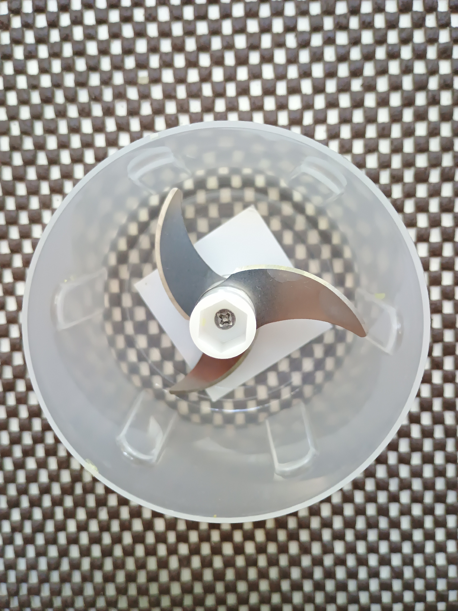 An overhead shot shows a blender blade assembly inside a clear plastic container. The container is placed on a brown and white checkered mat. The blender blade consists of three silver, curved blades attached to a white hexagonal center. The container and blade are positioned in the center of the frame. The background is a repeating pattern of brown and white squares.