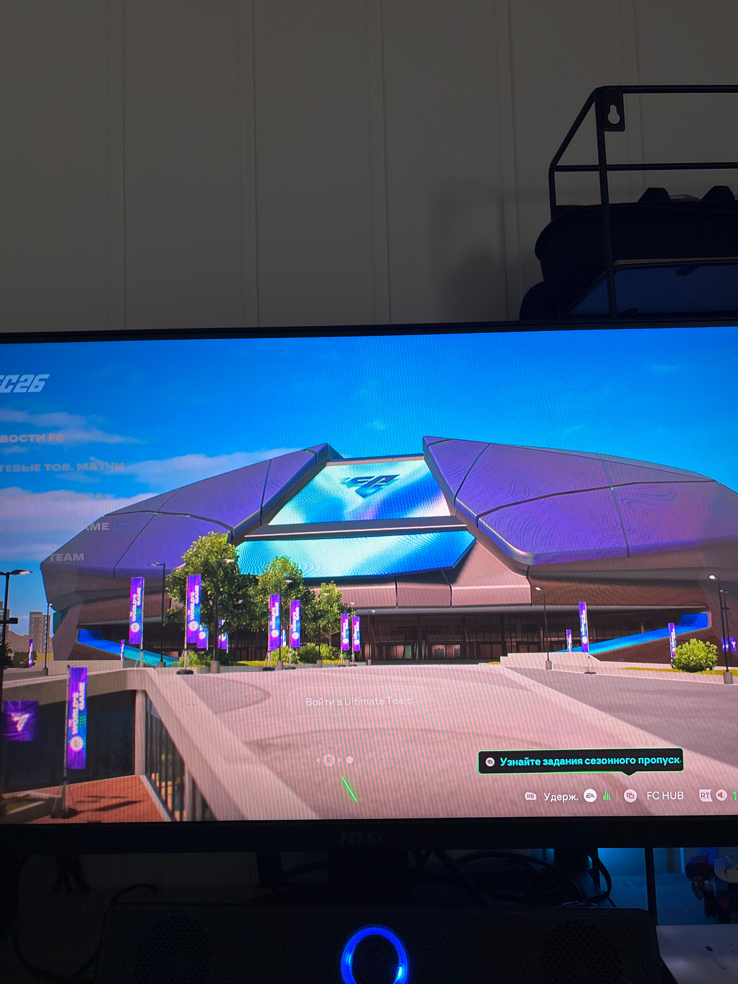 The image displays a video game interface, likely a sports simulation, featuring a futuristic stadium. The screen shows a vibrant blue sky and a large, architecturally modern stadium with purple and blue accents. Banners with purple designs are visible in front of the stadium. Text overlays on the screen indicate game options and notifications, including "FC26", "НОВОСТИ РФ", "Узнайте задания сезонного пропуска", and controller button prompts. The lower portion of the image shows the edge of a monitor or television screen, with some ambient lighting reflecting off its surface. The setting is clearly digital and does not depict any real-world location such as Isdalstø, Norway. There are no people, animals, or any objects from the physical world depicted in the image beyond the display device itself.