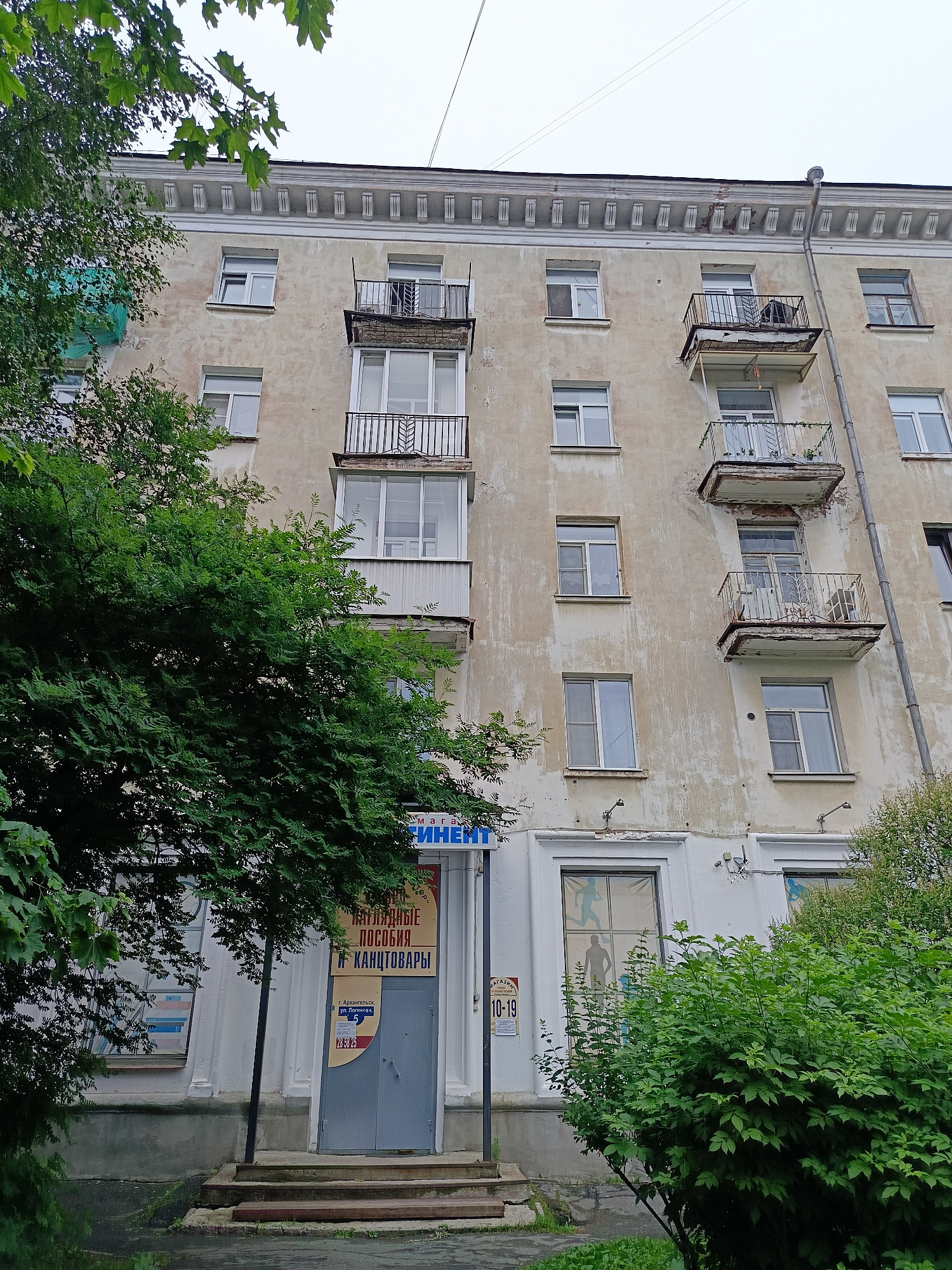 The image captures a multi-story building in Arkhangelsk, Russia, likely an apartment building given its architectural style and the presence of balconies. The building's facade is weathered and shows signs of age, with peeling paint and stained walls. Several windows are visible, some with their blinds or curtains drawn. Balconies, some with rusted railings, jut out from the upper floors. Lush green trees and bushes frame the lower part of the building, with one large tree partially obscuring the left side of the facade. A storefront is visible at the ground level, with a blue sign that has Cyrillic text and appears to be for a shop selling stationery and office supplies. A gray door with a sign displaying hours of operation is also part of the storefront. The sky is overcast, suggesting a cloudy day. There are no people visibly present in the image.