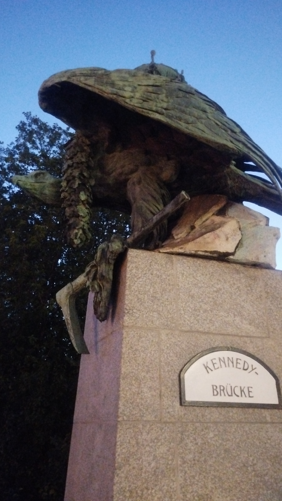 The image shows a statue of an eagle on a granite pedestal. The eagle is made of bronze and is weathered green. It has large wings spread and is holding a bundle of leaves in its talons. The pedestal has a plaque with the words "KENNEDY BRÜCKE" inscribed on it. The statue is outdoors, with a dark sky in the background and trees visible to the left. The lighting suggests it is either dusk or dawn. The overall impression is one of a monument or memorial.