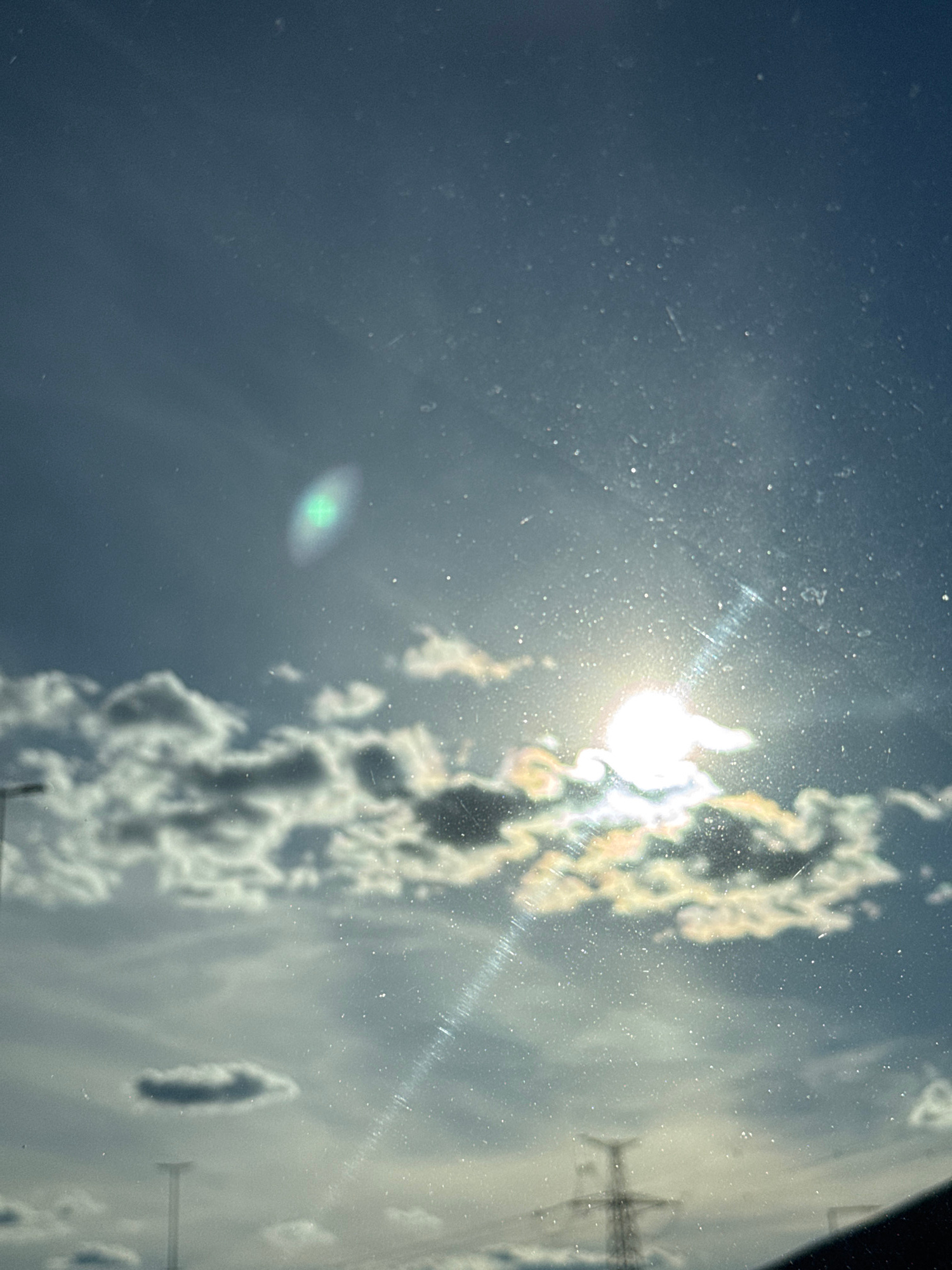 A cloudy sky with the sun shining brightly through the clouds. There is a bright green light on the left side of the image, and a rainbow effect on the right side. In the foreground, there are some power lines and a pylon. The image is slightly blurry and there are some specks on the lens.