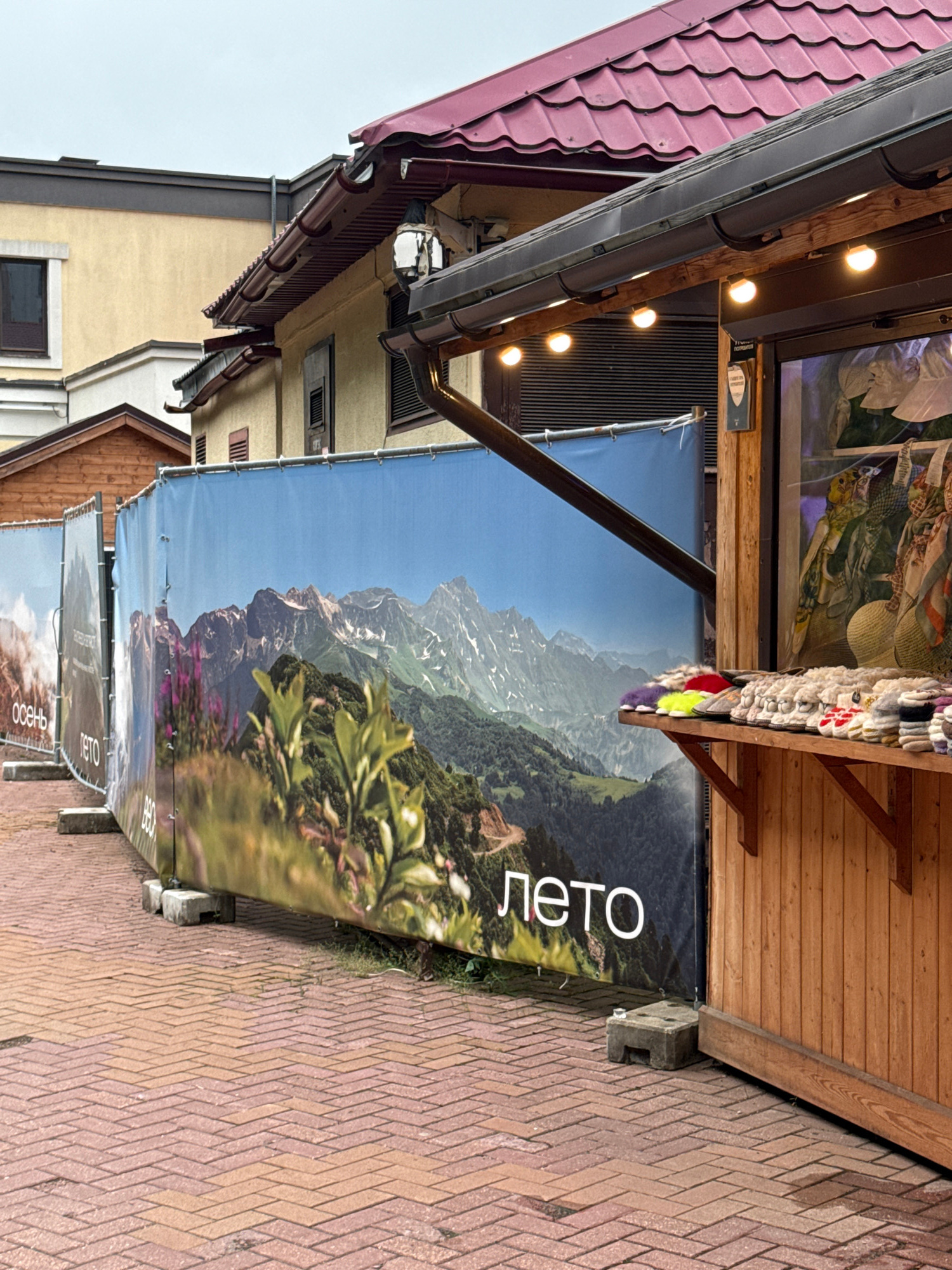 The image depicts an outdoor scene in Estosadok, Russia, likely a market or a shopfront. A large banner with a mountainous landscape and the Russian word "лето" (summer) is prominently displayed. Below this, another banner with the Russian word "осень" (autumn) is partially visible. These banners suggest a seasonal display or sale.
The setting includes a wooden structure with a red-tiled roof, suggesting a building or an awning. Several string lights are illuminated, indicating it might be late afternoon or evening, or simply for ambient lighting. The ground is paved with brick-like stones in a diamond pattern. Adjacent to the paved area, there is some green foliage and grass.
The overall impression is one of a commercial area, possibly in a tourist destination given the scenic imagery on the banners. There are no people clearly visible in the frame. The weather appears to be overcast. The focus is on the banners and the rustic architectural elements of the stall.
