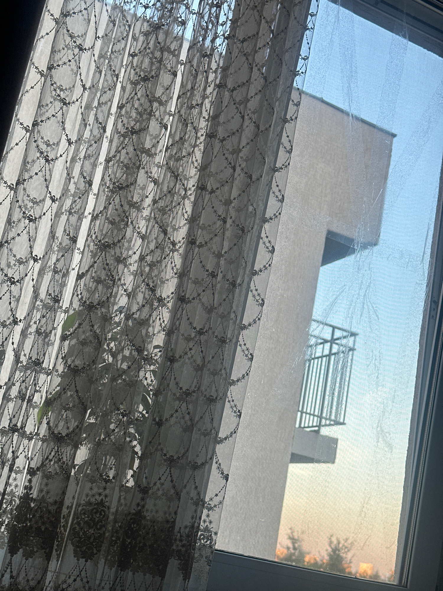 A sheer, patterned curtain is partially drawn across a window, obscuring the view of a modern building outside. The curtain has a delicate, intricate design of small floral motifs and star-like shapes, arranged in a repeating pattern that creates a scalloped edge along its bottom and sides. The sunlight filters through the curtain, casting soft shadows and highlighting the texture of the fabric. Beyond the curtain, a portion of a building is visible, characterized by its clean lines and a contemporary architectural style. A small balcony with a metal railing juts out from the building's facade. The sky outside appears clear, with a gradient of blue hues suggesting either dawn or dusk. In the lower part of the window, a faint outline of trees and distant buildings can be seen, illuminated by the warm light of the setting or rising sun. The windowpane itself has some smudges and streaks, which are faintly visible against the outdoor light.
